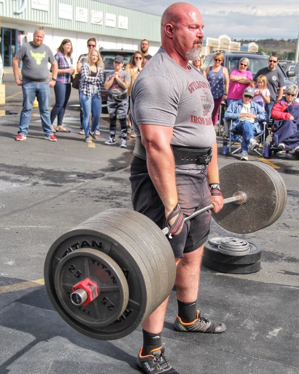 Man deadlifting a barbell with heavy weights outdoors, observed by a crowd.