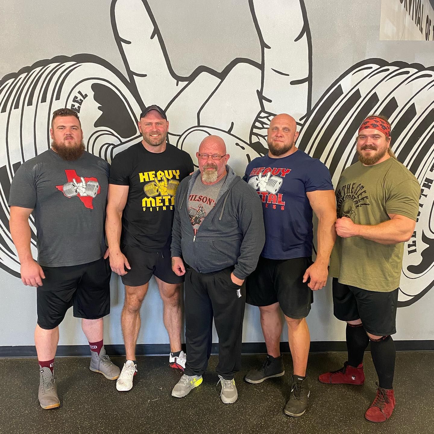 Five strongmen pose in a gym; a large peace sign and barbell art are behind them.