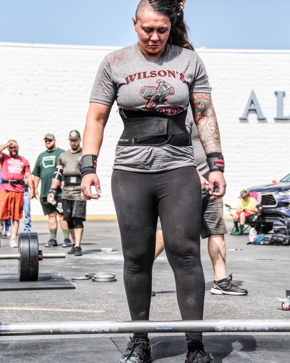 Woman wearing lifting belt, prepares to lift a barbell outdoors. Other people are in the background, near a building.