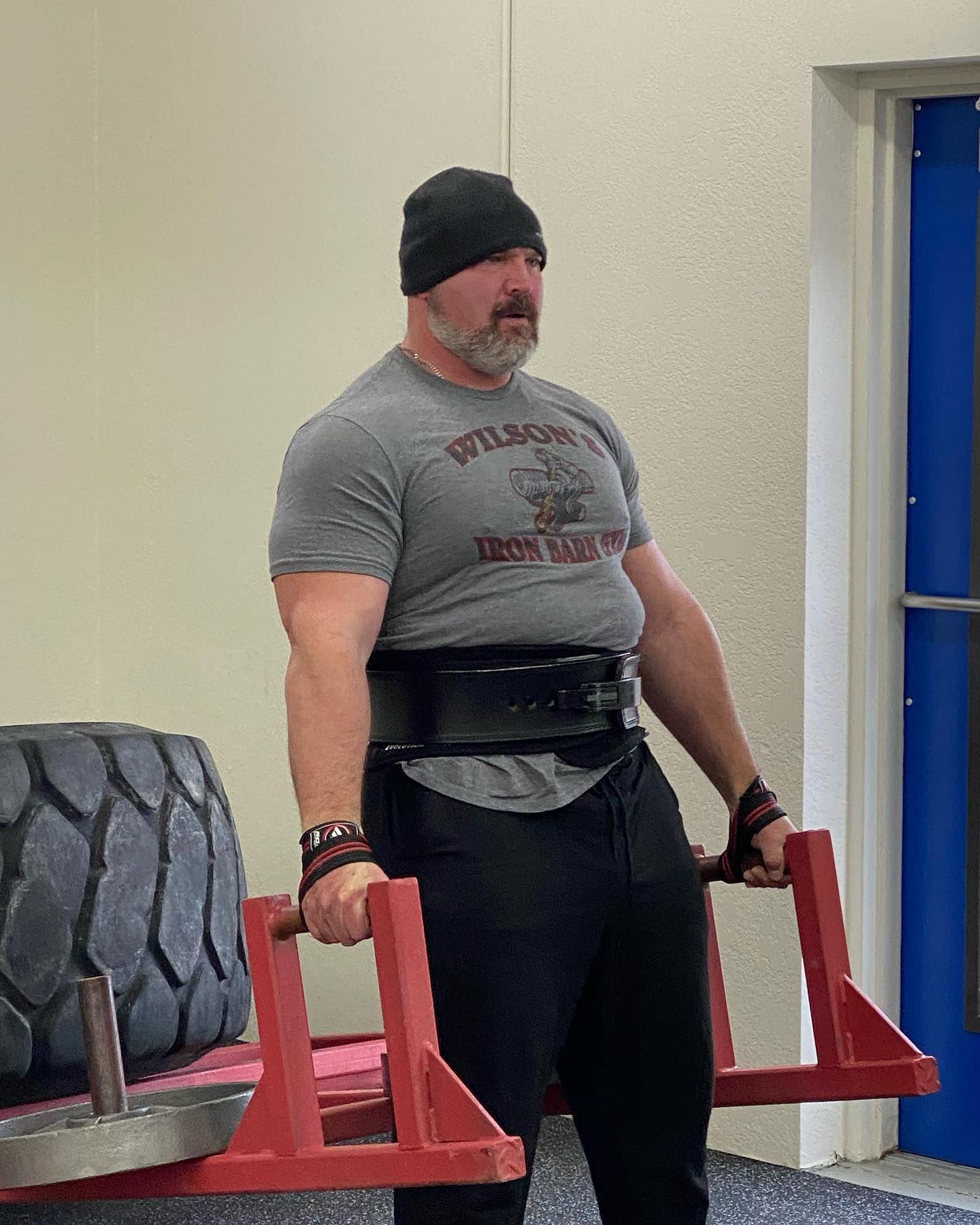 Man lifting weights, wearing a black beanie, weight belt, and wrist wraps, with a tire and frame in a gym.