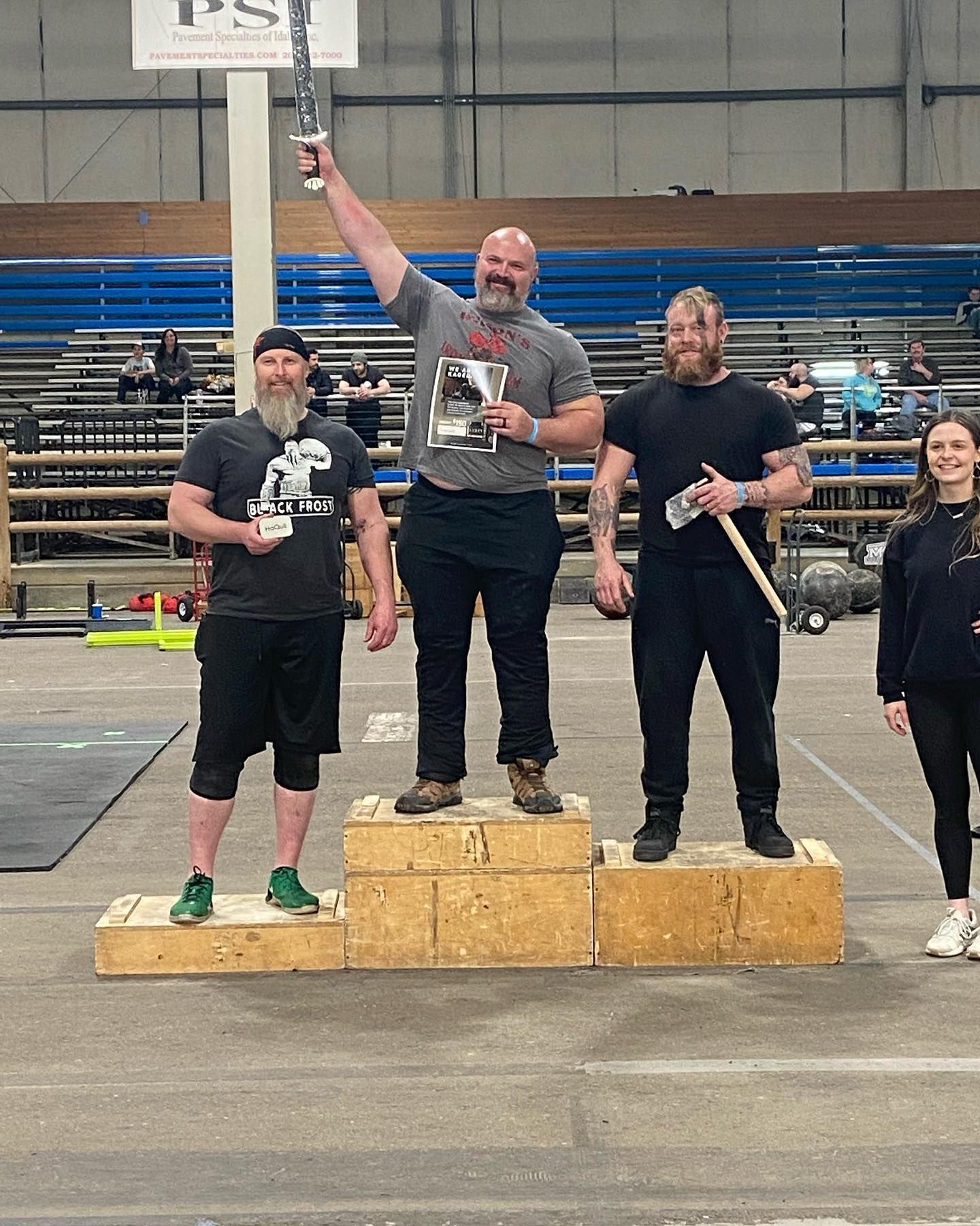 Strongman competition winners on podium; man in center holds trophy, smiles and raises arm. Two others stand nearby.