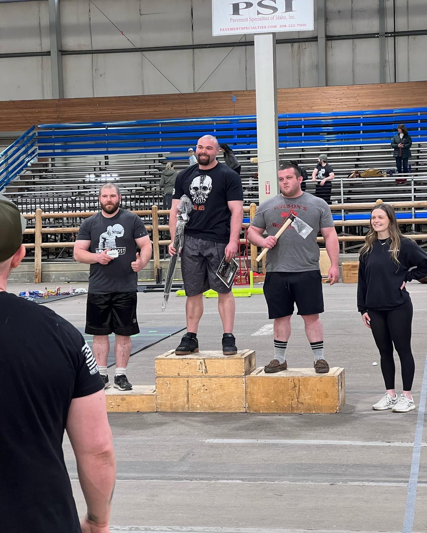 Podium of strongmen: man with skull shirt on top, others with axes, indoor competition setting.