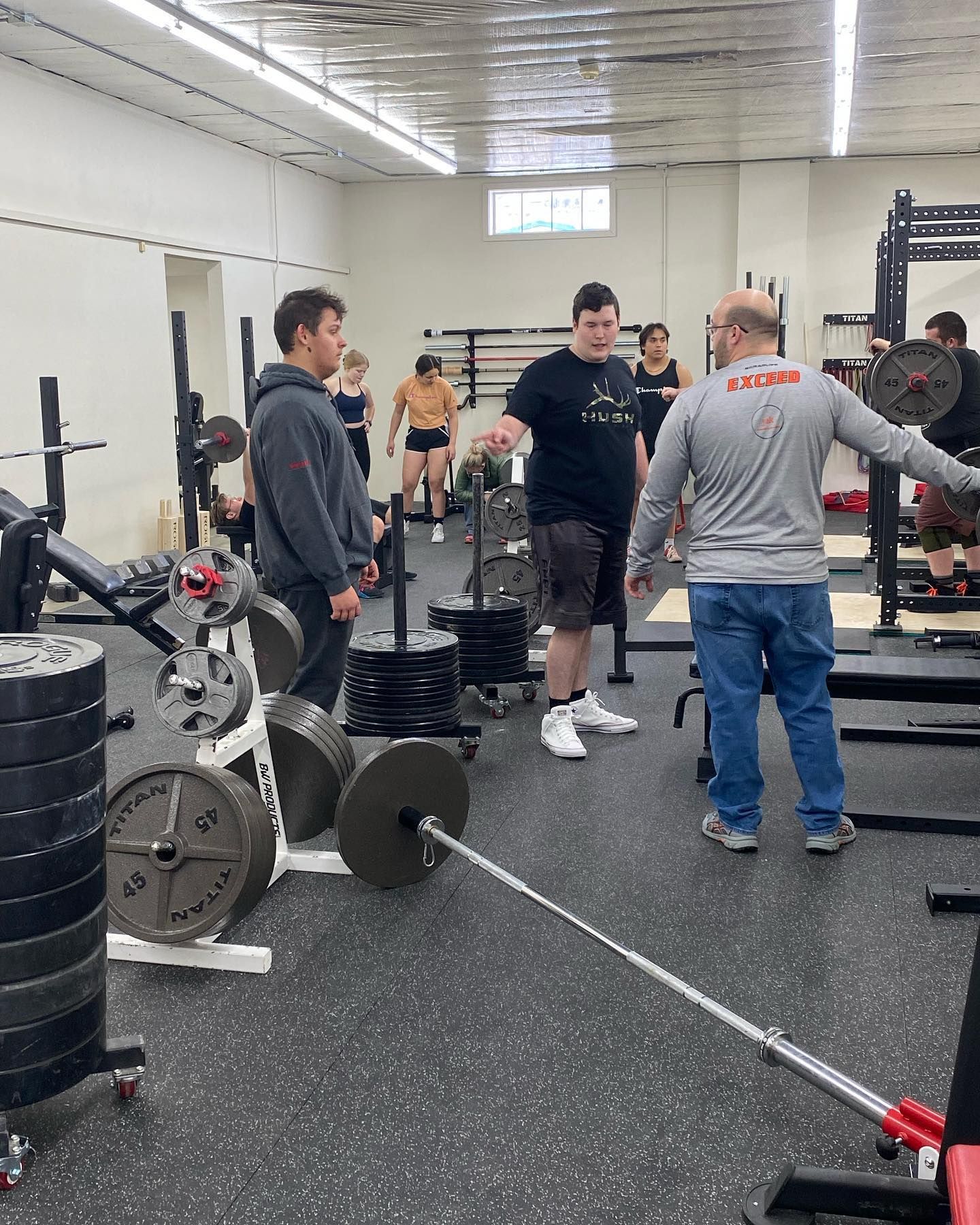 People lifting weights in a gym with barbells, weight plates, and a trainer observing.