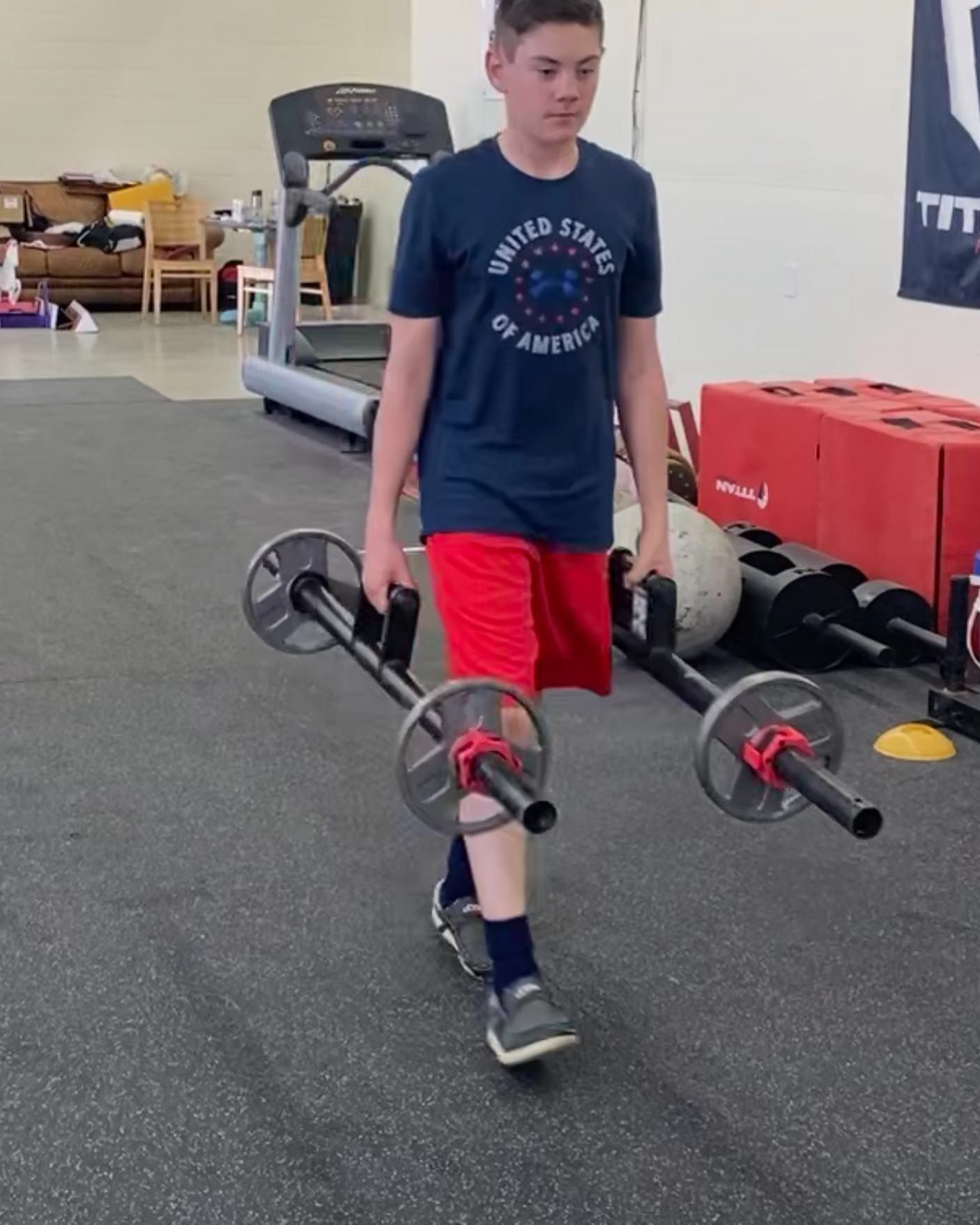 A person in a gym carrying two barbells, walking. Wearing a blue t-shirt and red shorts.
