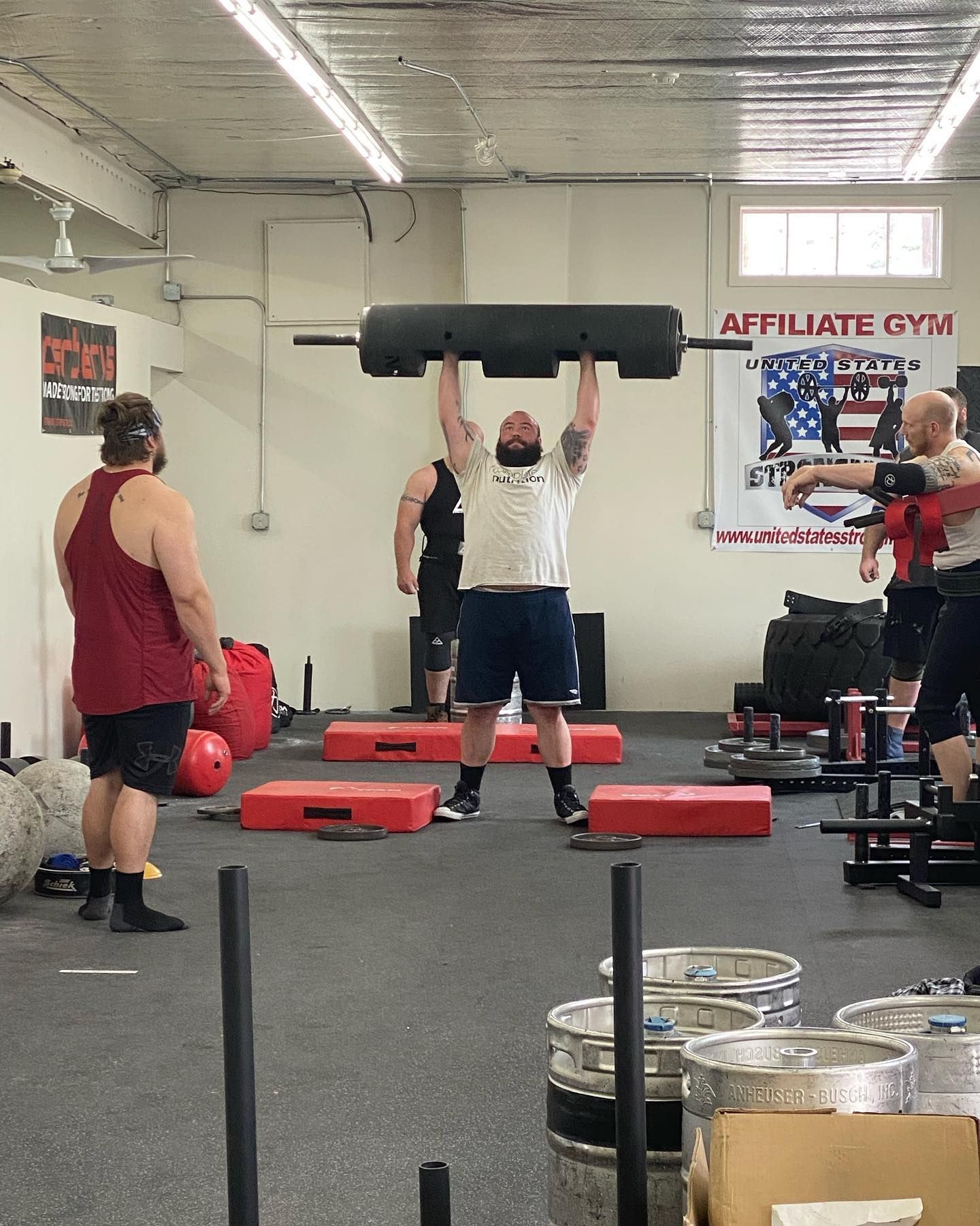 Man lifts a heavy log overhead at an indoor gym. Other men watch and spot.