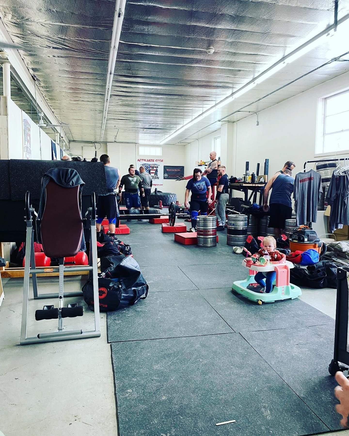 People lifting weights in a gym. A baby in a walker is nearby. Gray flooring, reflective ceiling.