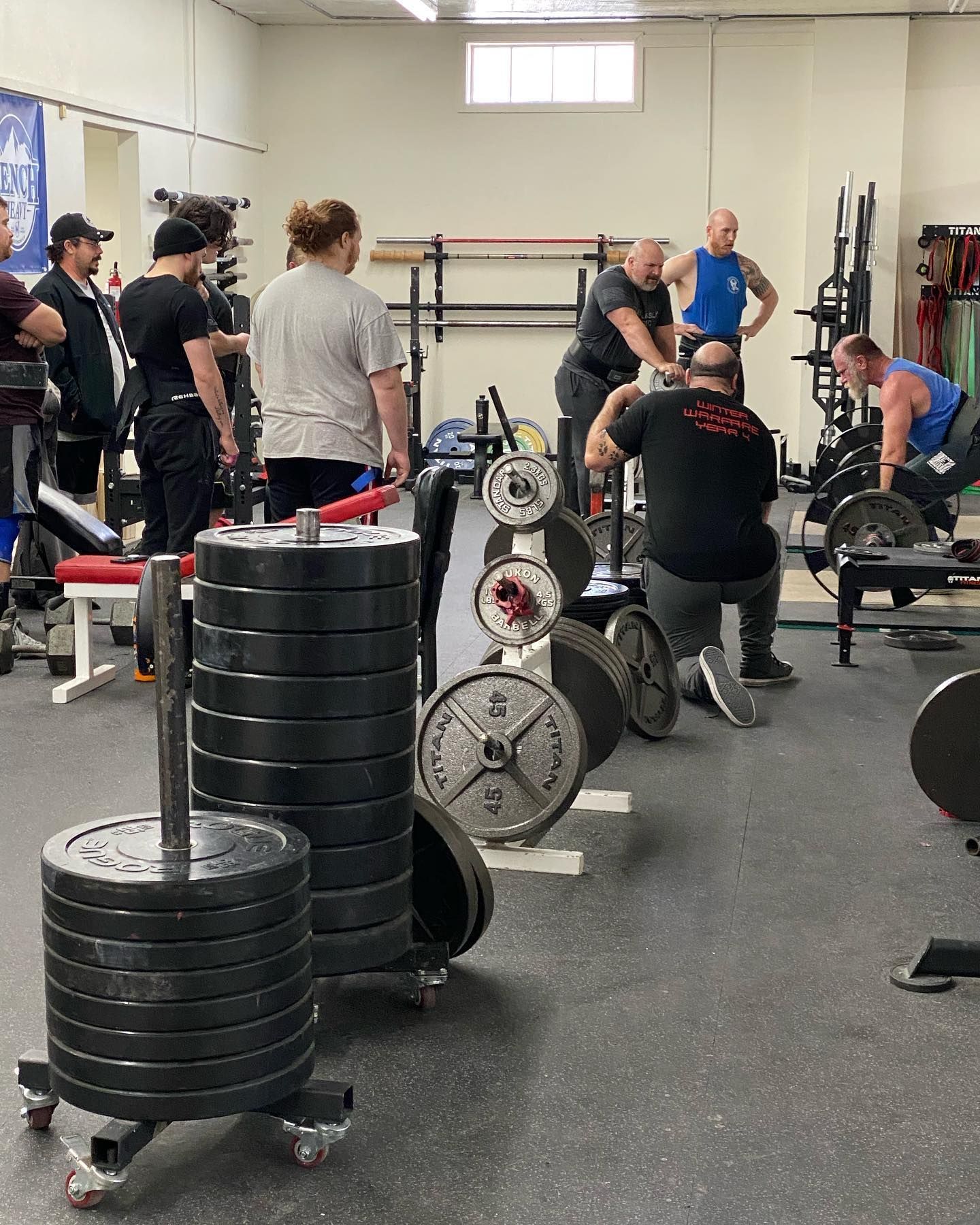 Weightlifters in a gym, some spotting, others prepping or lifting barbells. Gray and black weights stacked.