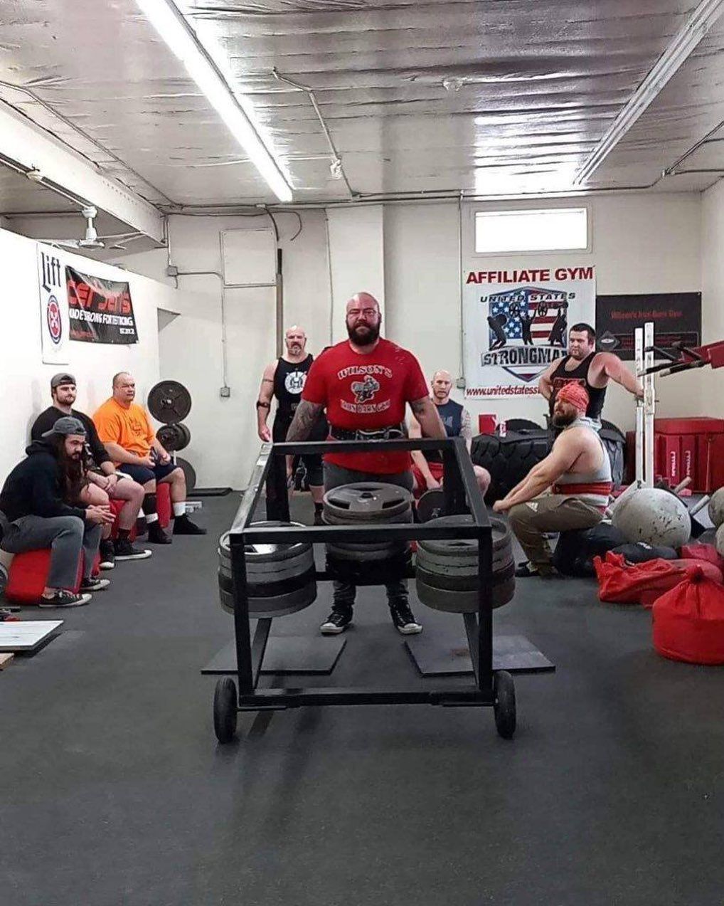 Strongman in red shirt lifts a heavy frame loaded with weights, observed by others in a gym setting.