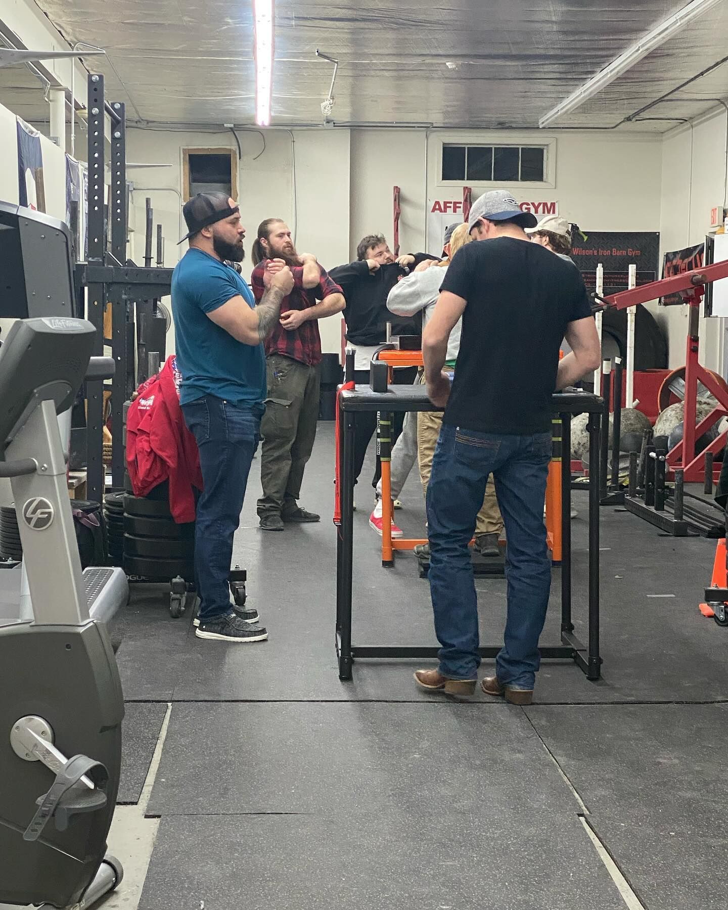People arm wrestling in a gym. Men are standing around a table, watching.