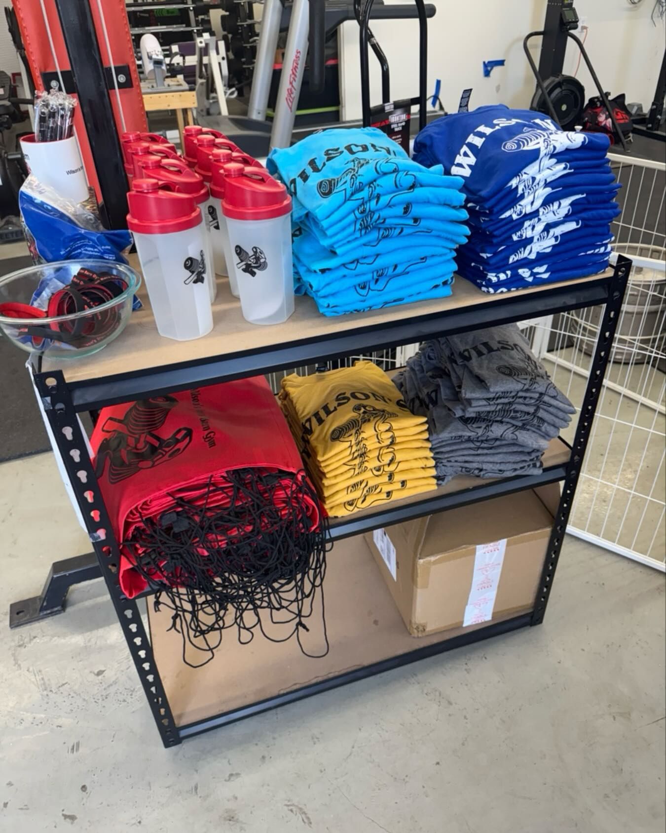 Shelving unit holding stacks of t-shirts, shaker bottles, and a box, likely for a gym or fitness business.