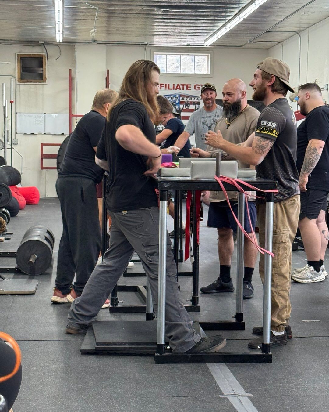 Arm wrestling match in a gym, two men at a table, others watching.