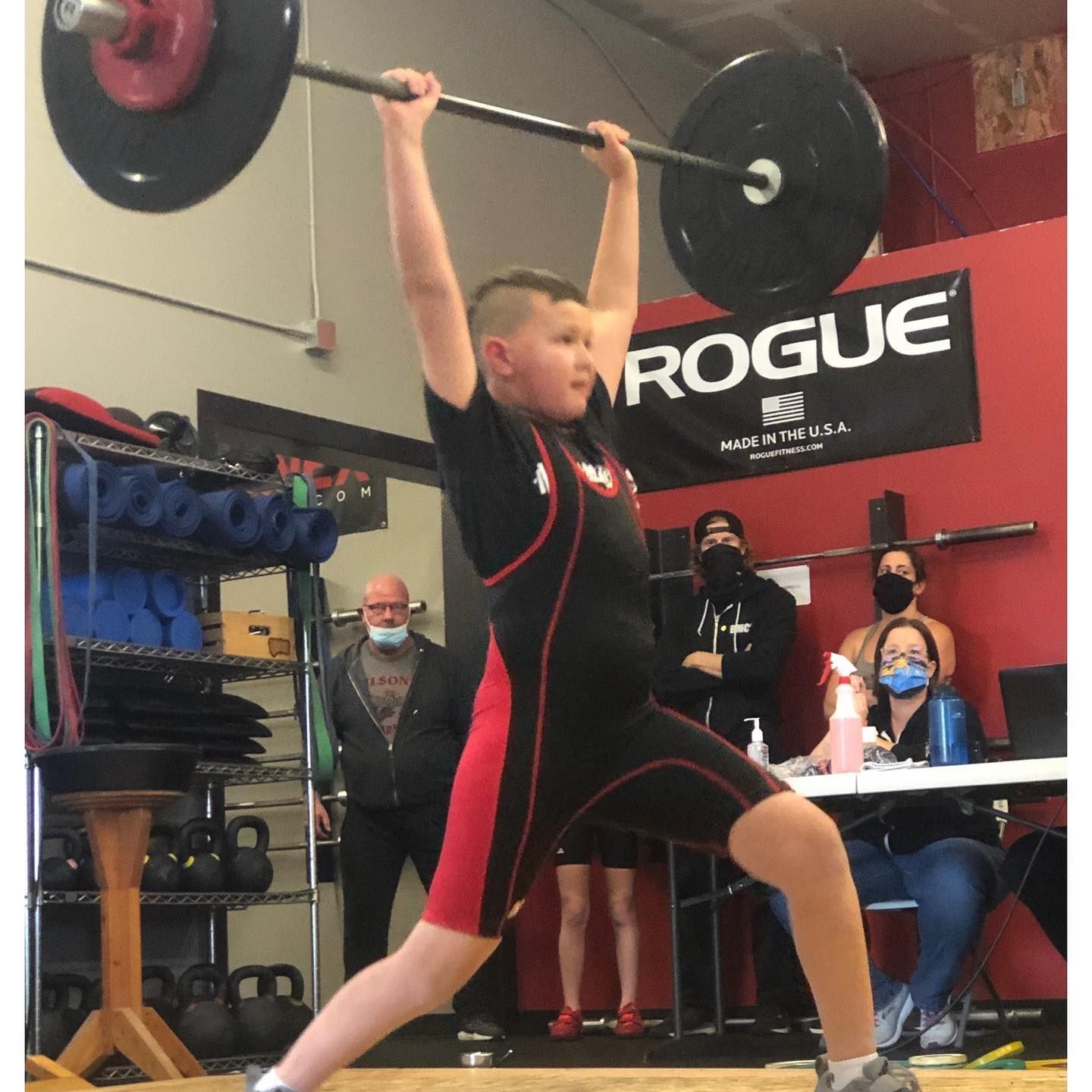 Boy weightlifting overhead in a gym, wearing a black and red uniform.