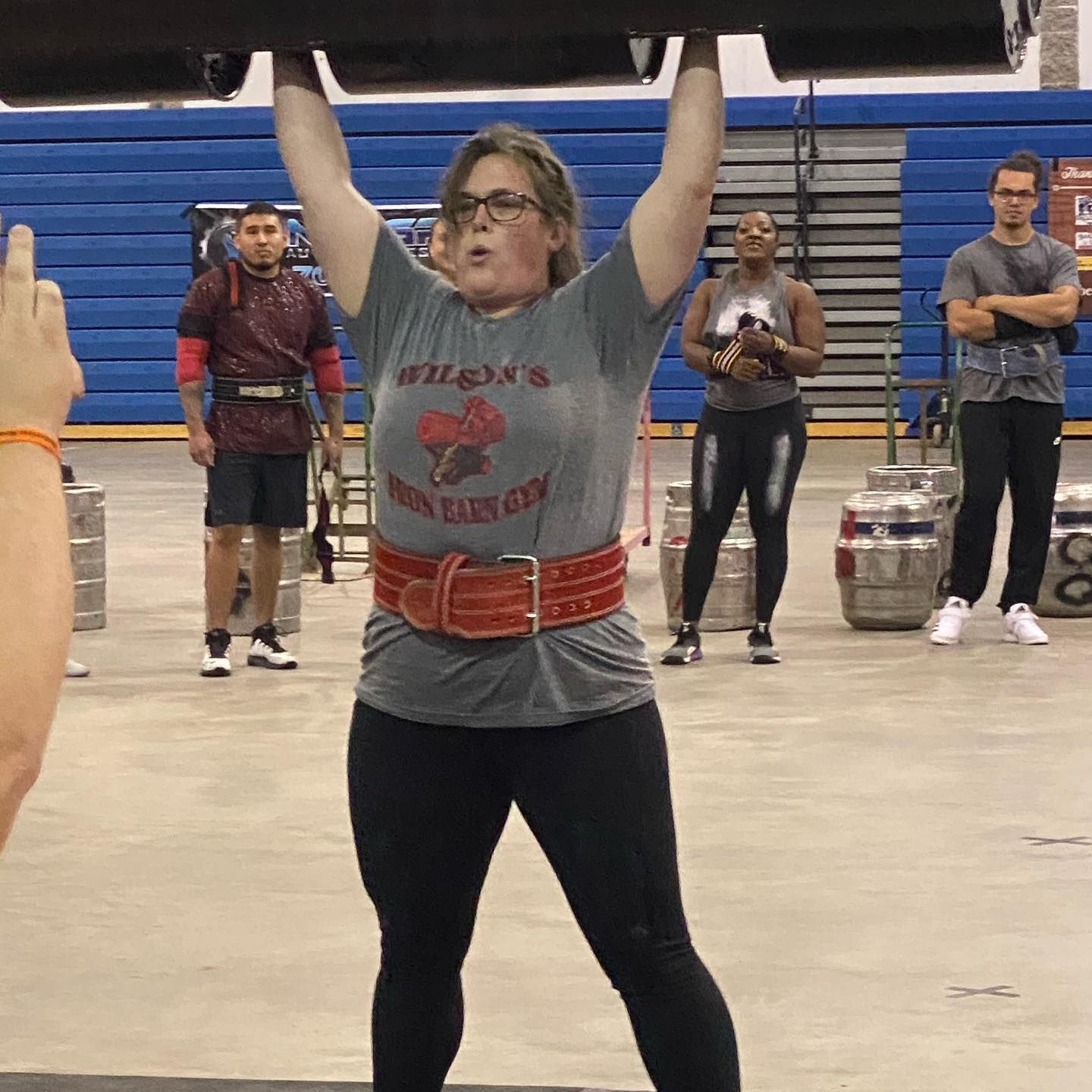 Woman lifting a heavy log overhead at a gym, wearing a weight belt and gym shirt.