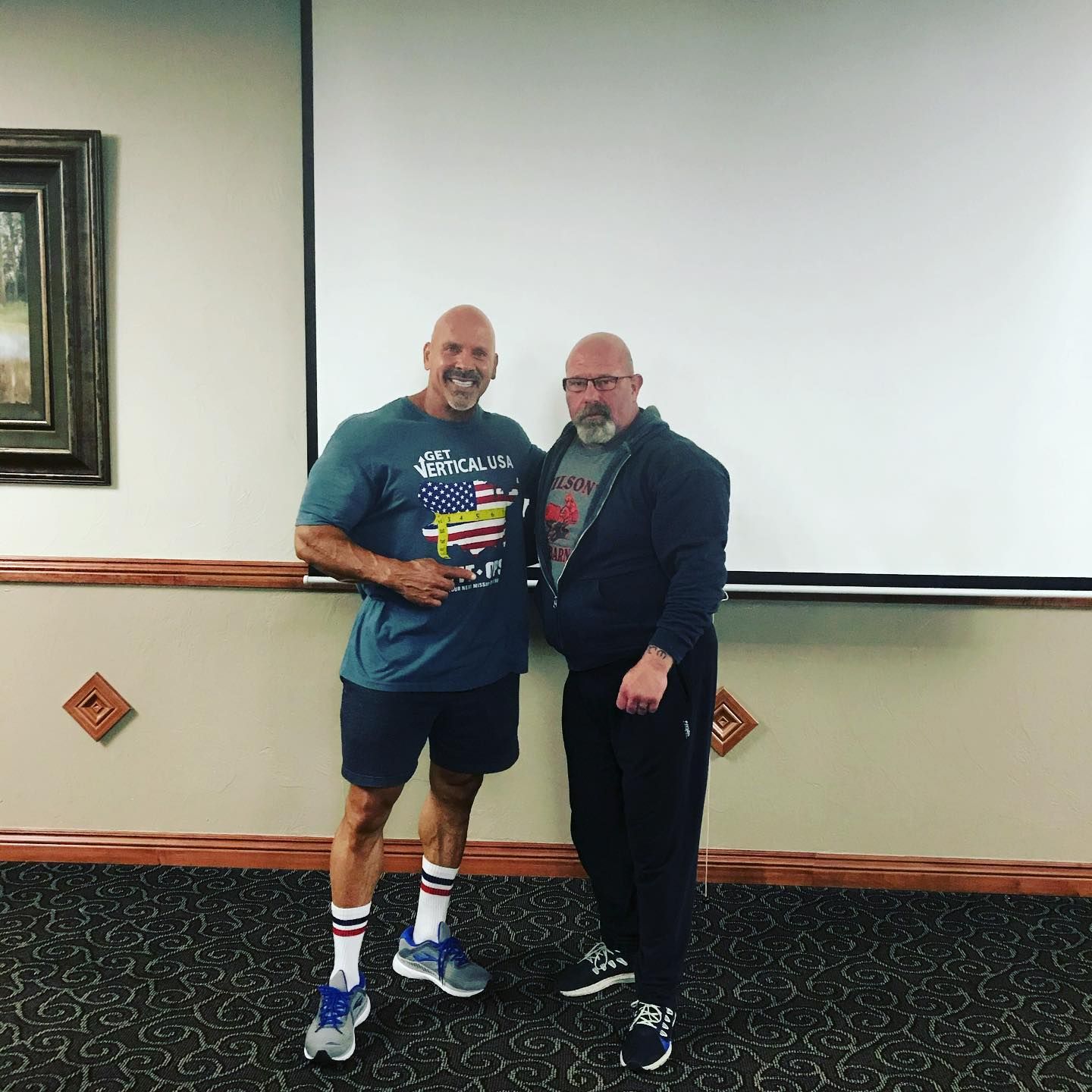 Two men stand smiling in front of a white screen. One wears a tee and shorts, the other a jacket and sweats.