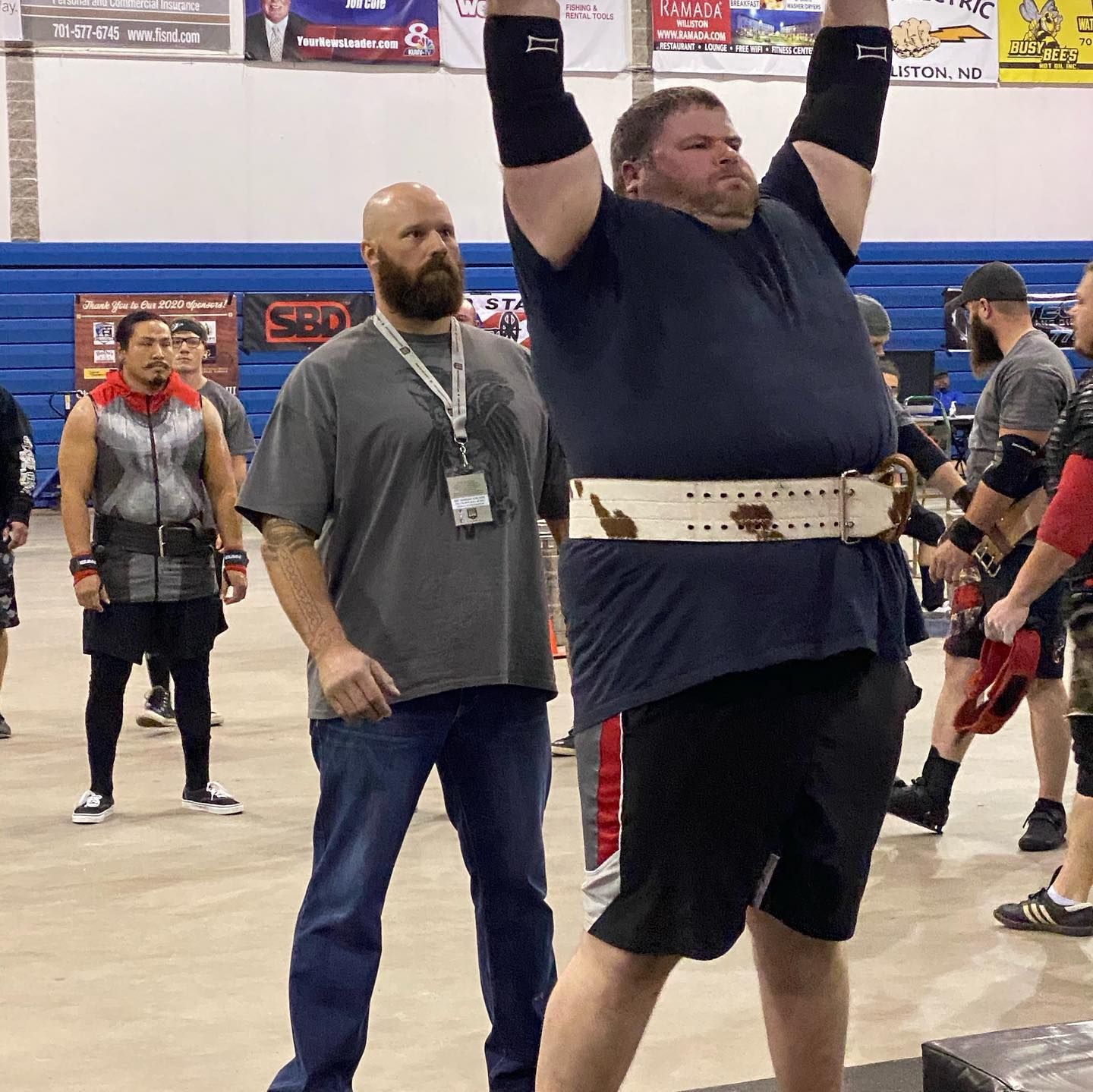Man in weightlifting belt overhead, coach observing. Indoor gym setting.