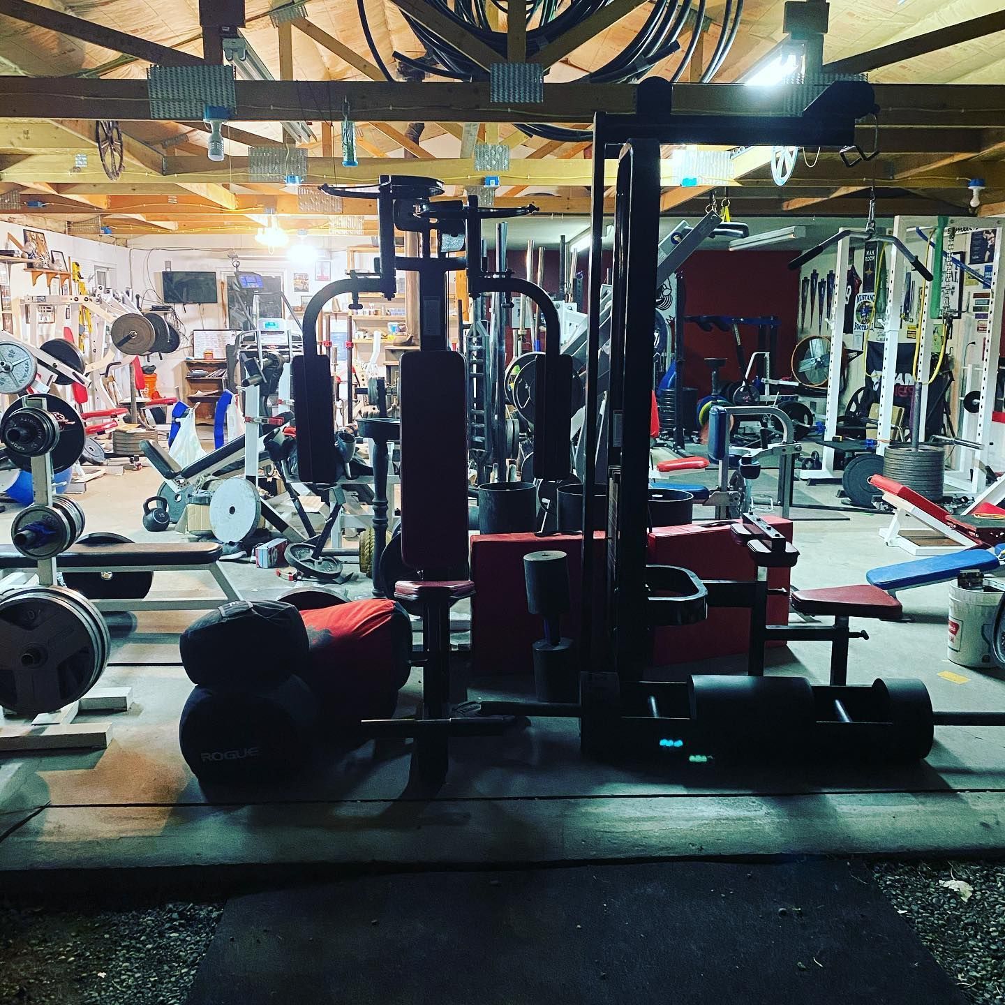 Weightlifting gym interior with various machines, weights, and tools. Low lighting and cluttered arrangement.