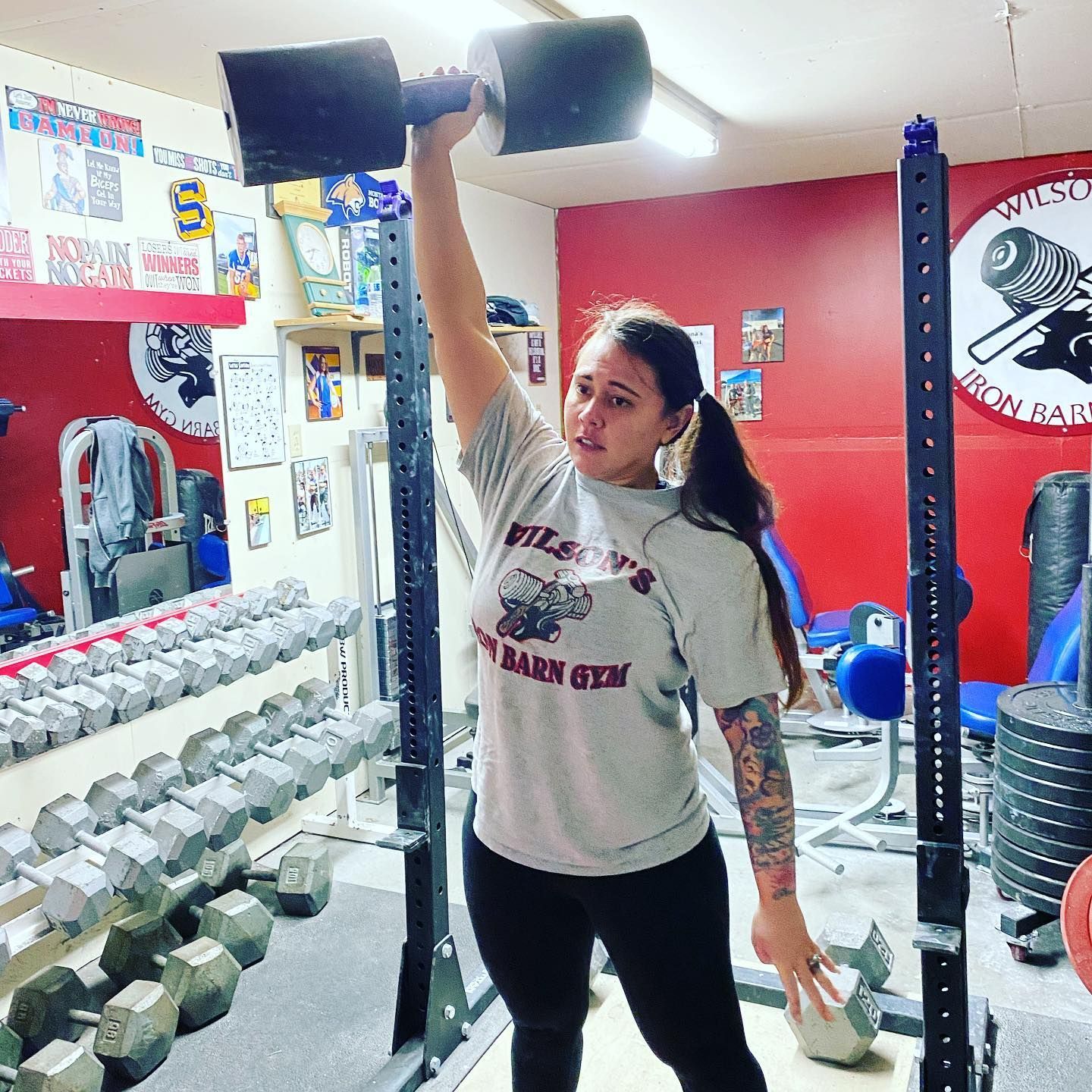 Woman lifting a large dumbbell overhead in a home gym. She wears a grey shirt and black leggings. Gym is red and metallic.