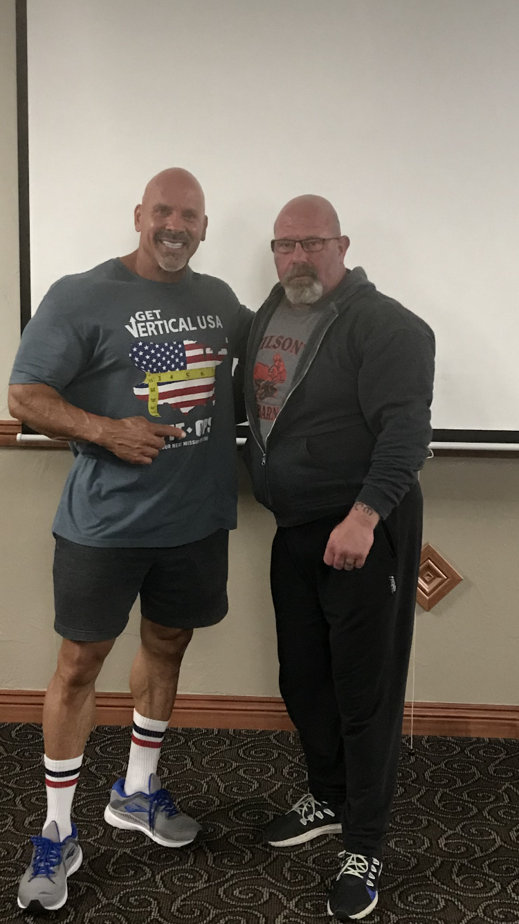 Two men posing for a photo indoors, one muscular in shorts and a tee, the other in a hoodie and sweatpants.