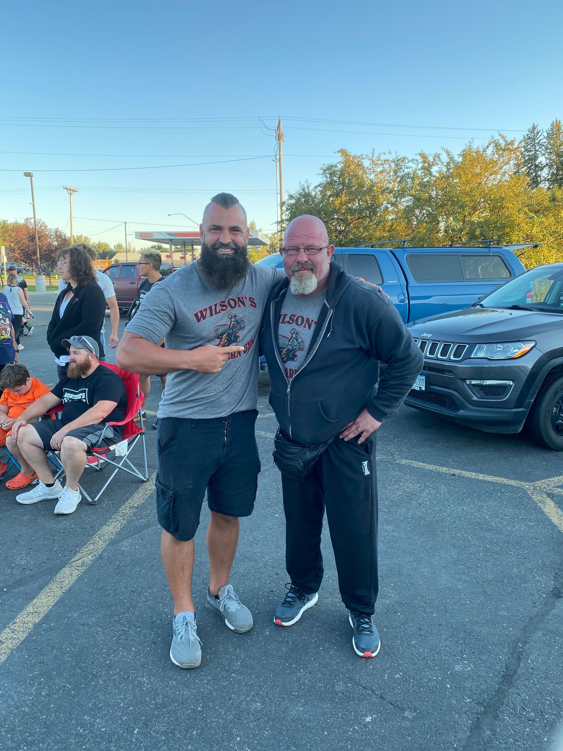 Two men pose outdoors. One points, the other smiles. Vehicles and people are in background.