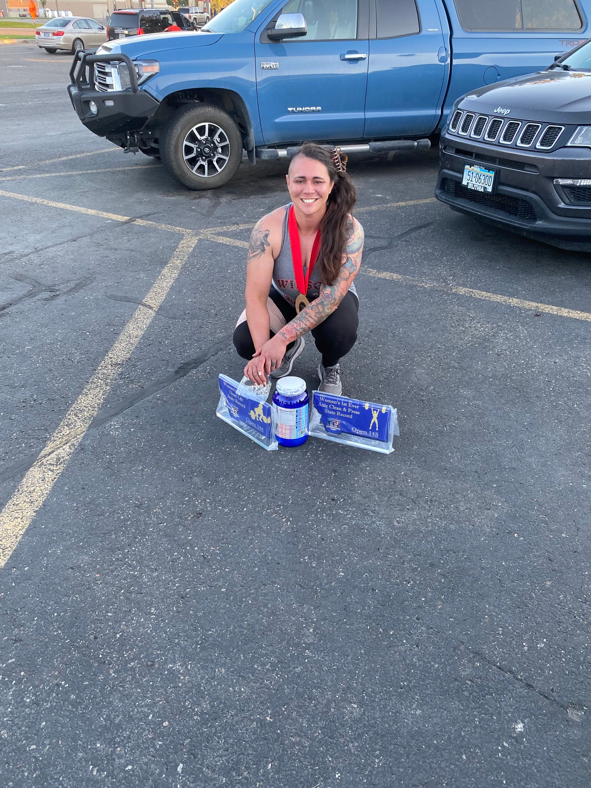 Woman kneels with trophy in a parking lot, wearing a medal. Blue truck and black SUV in the background.