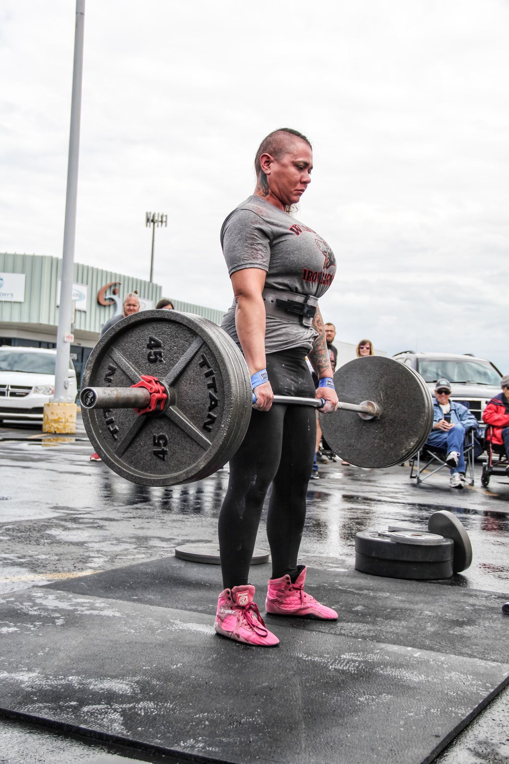 Woman lifting a barbell outdoors; wearing black leggings, gray shirt, and pink shoes.
