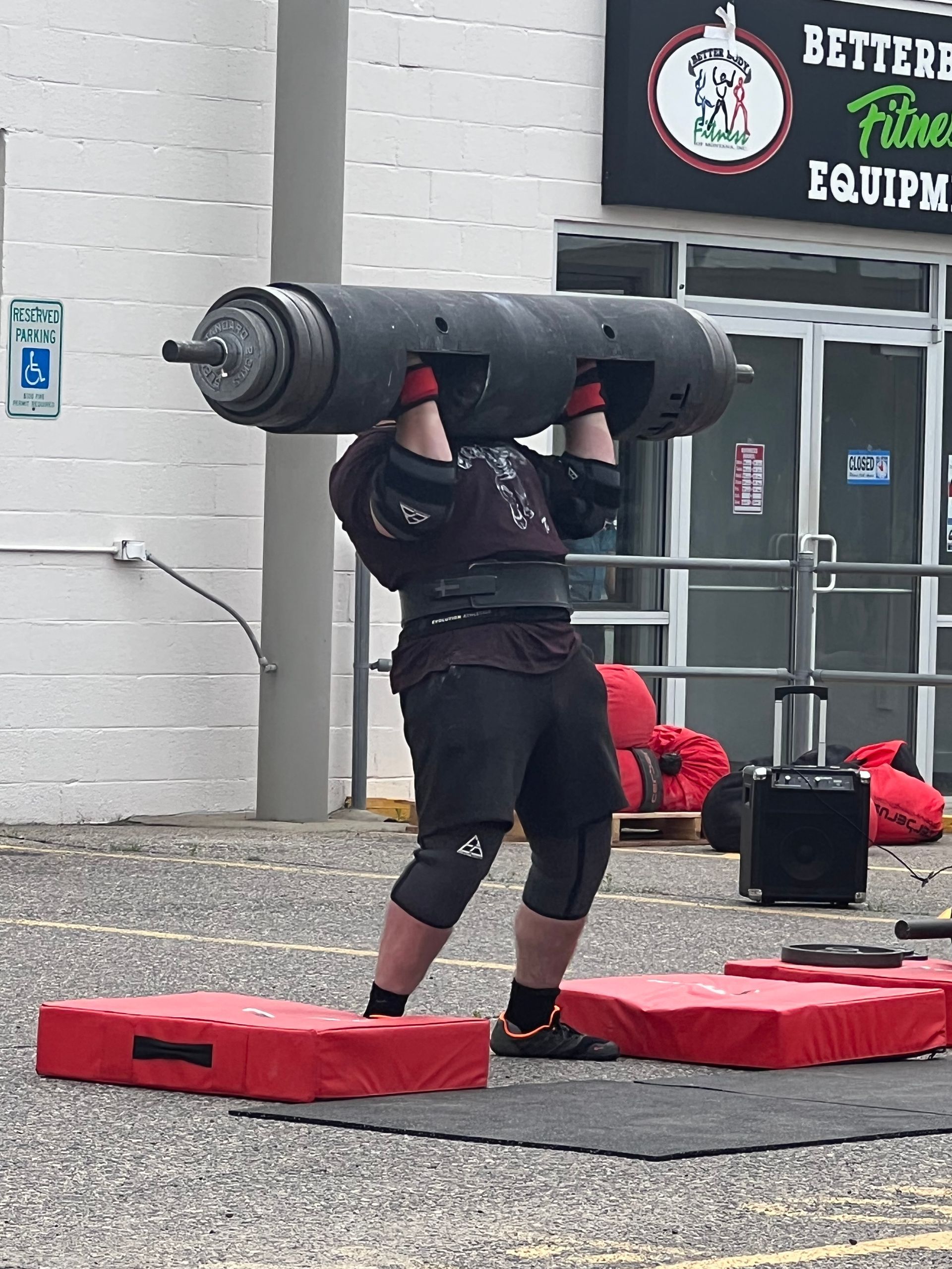 Man lifting a heavy log overhead during a strongman competition outside a fitness equipment store.