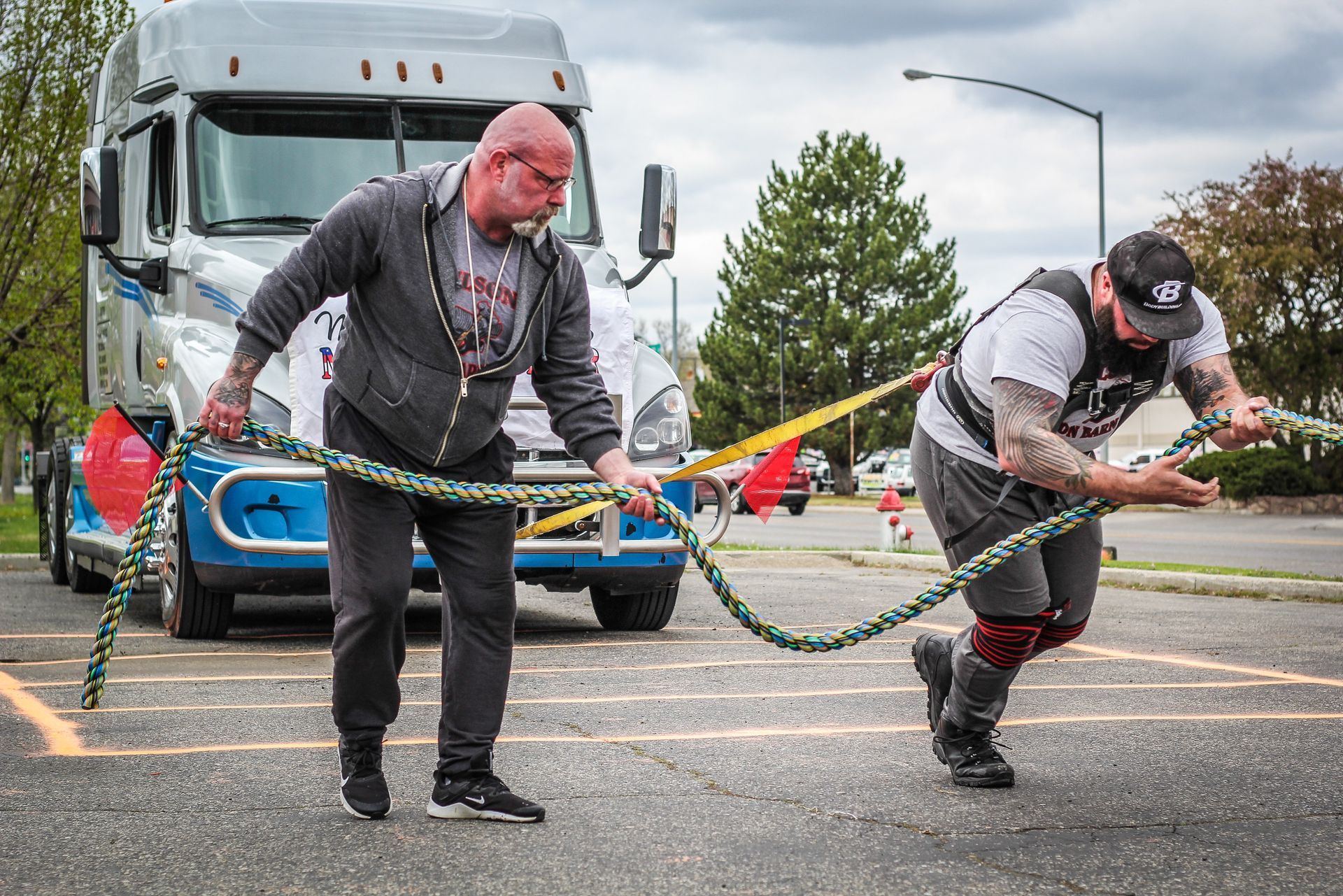 Two men pulling a large truck with a thick rope outdoors on a parking lot.