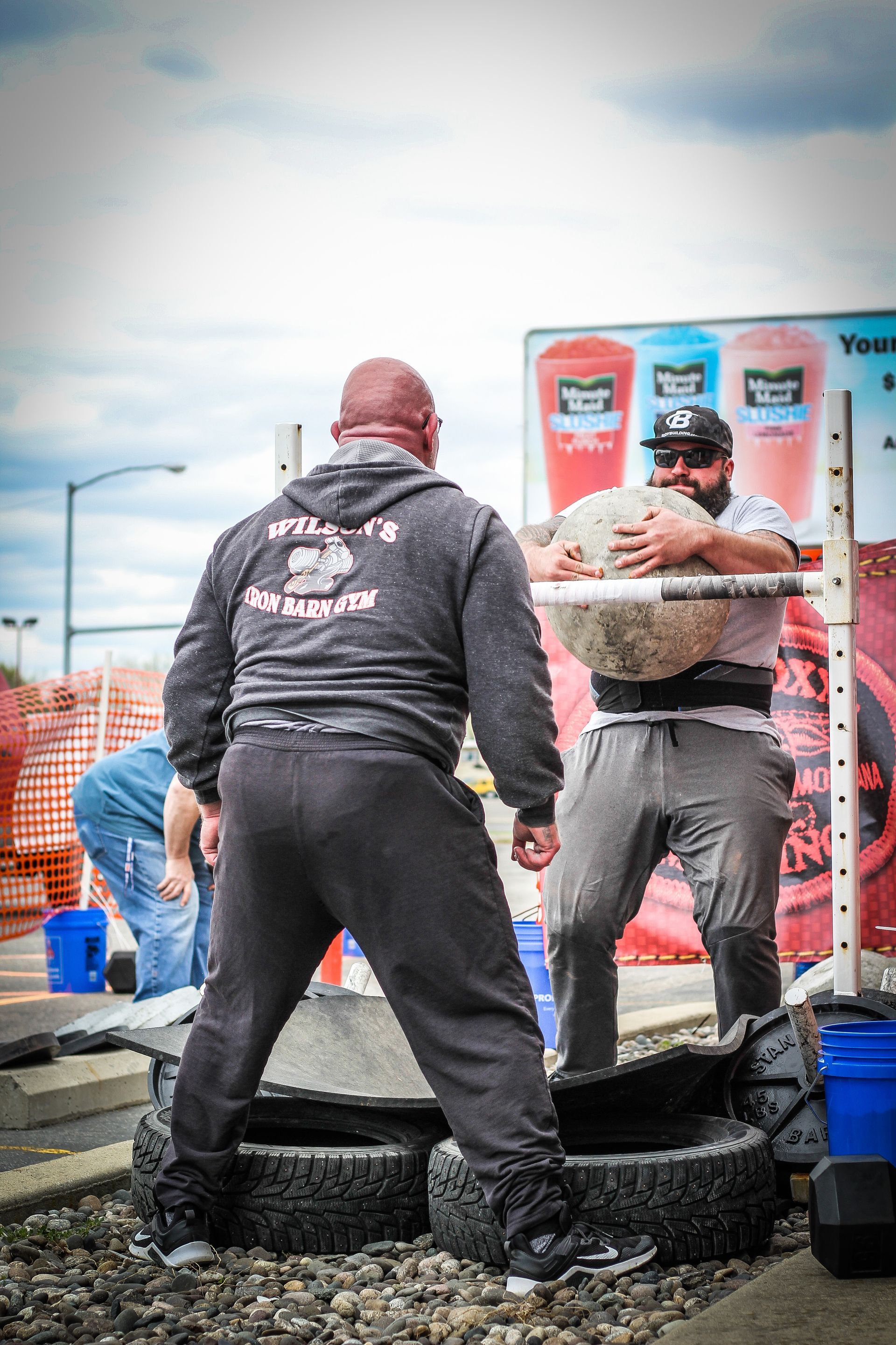 Strongman lifts a stone onto a platform made of tires; another man watches. Outdoor competition.