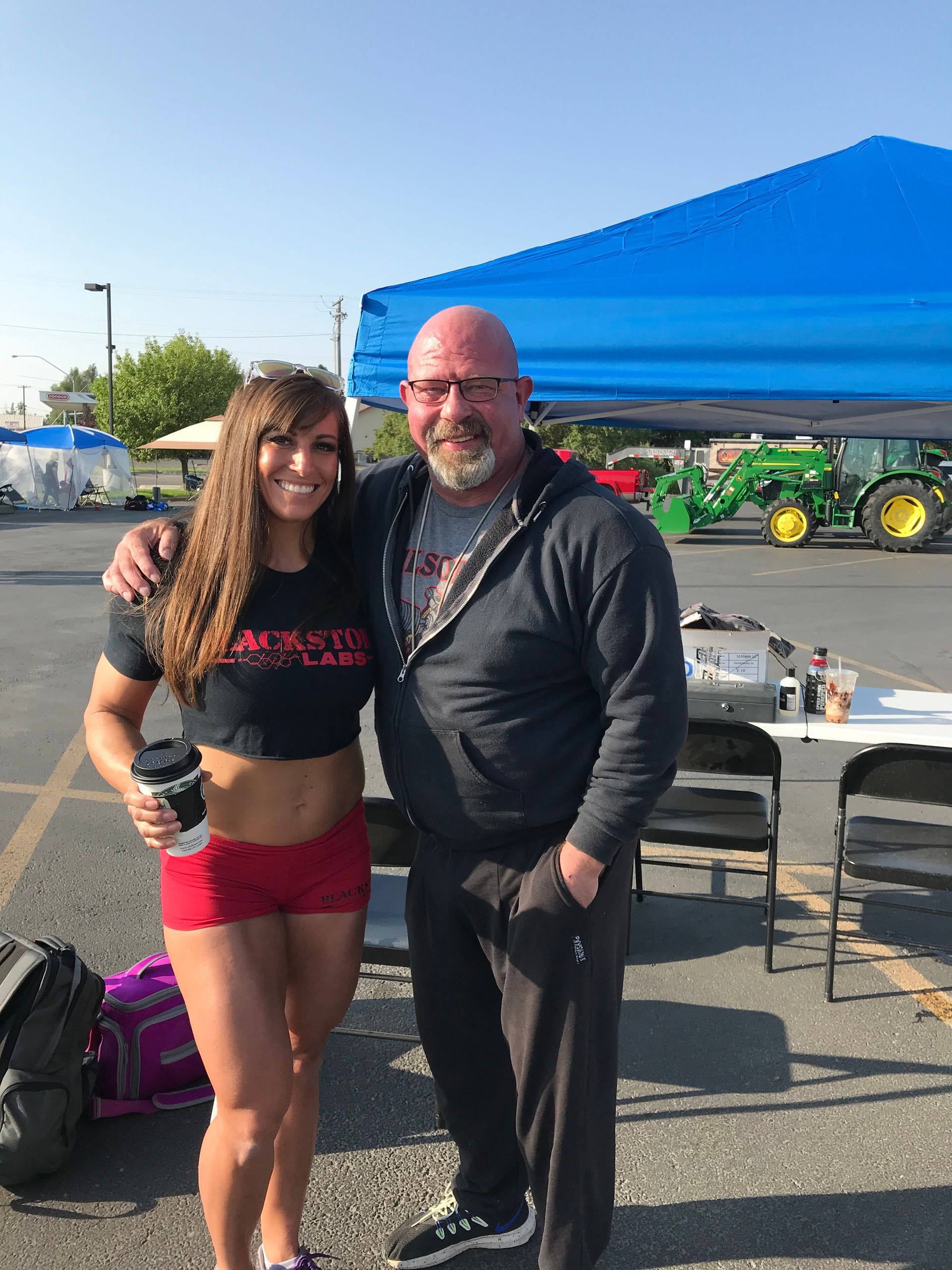 Woman and man posing outdoors, woman holding coffee, man's arm around her. Blue tent, tractor in the background.