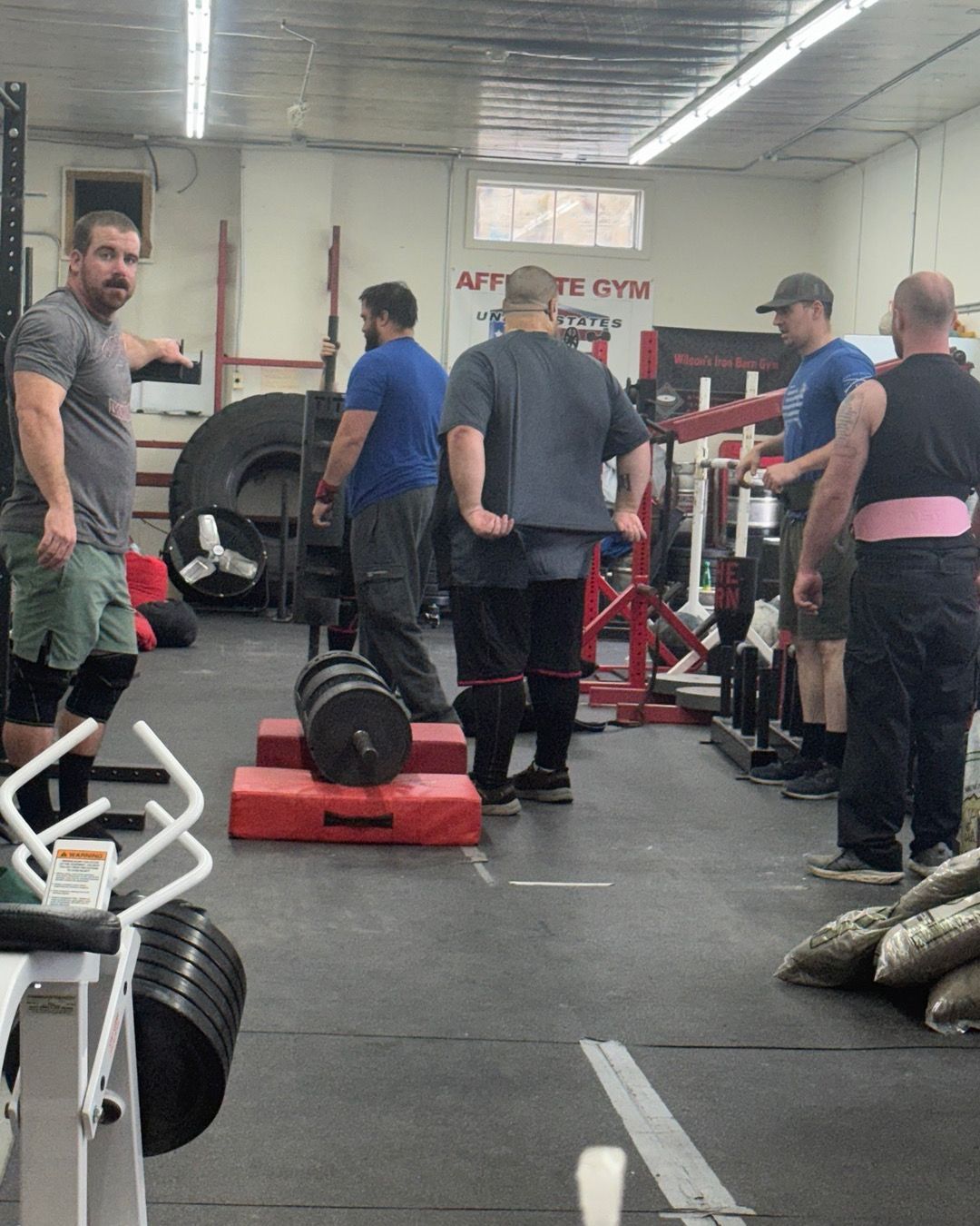 People lifting weights in a gym. A person in grey shirt points. Barbell on a platform. Other gym equipment present.