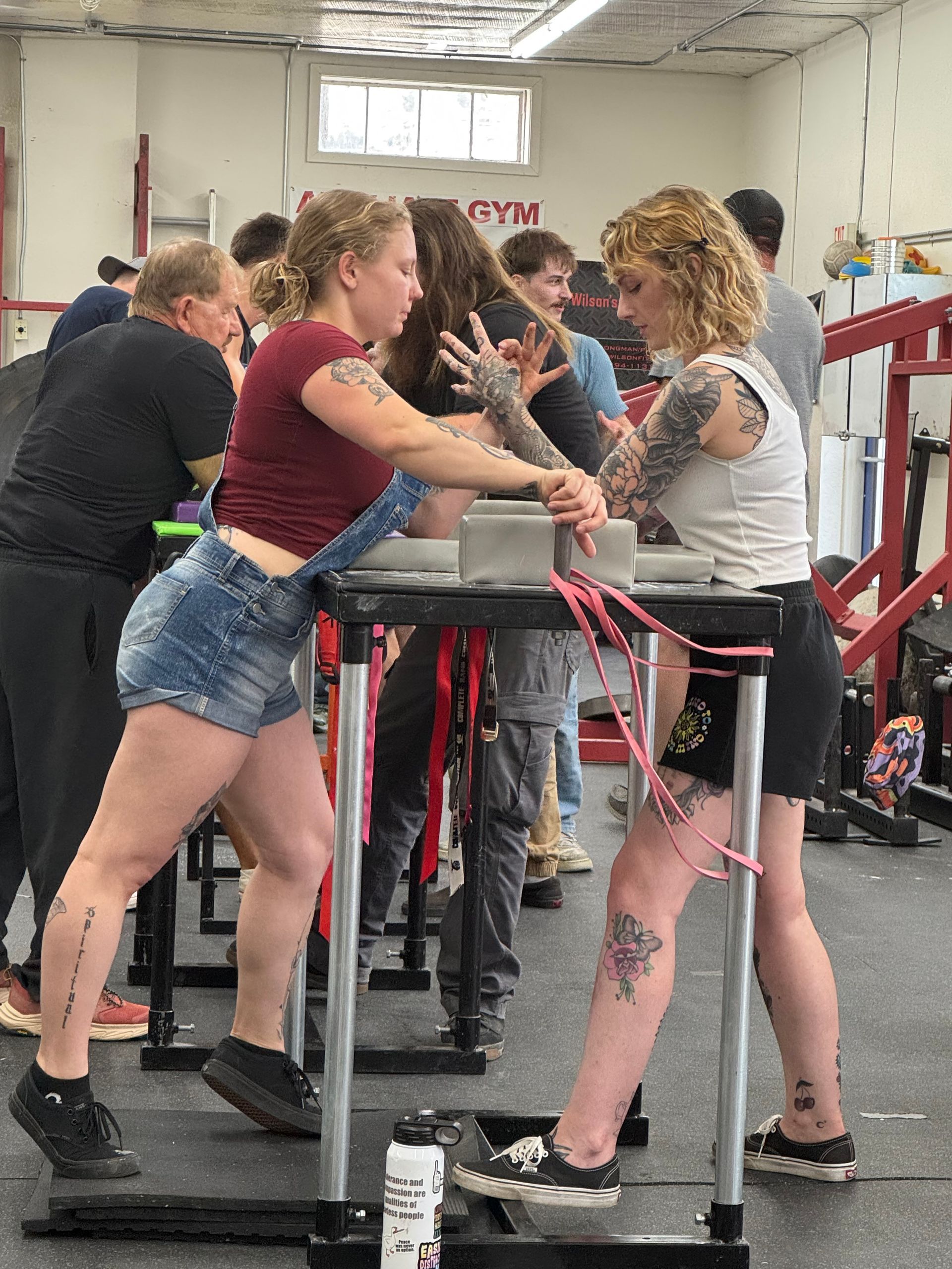Two women arm wrestling at a gym. One in overalls, the other in a tank top, surrounded by onlookers.
