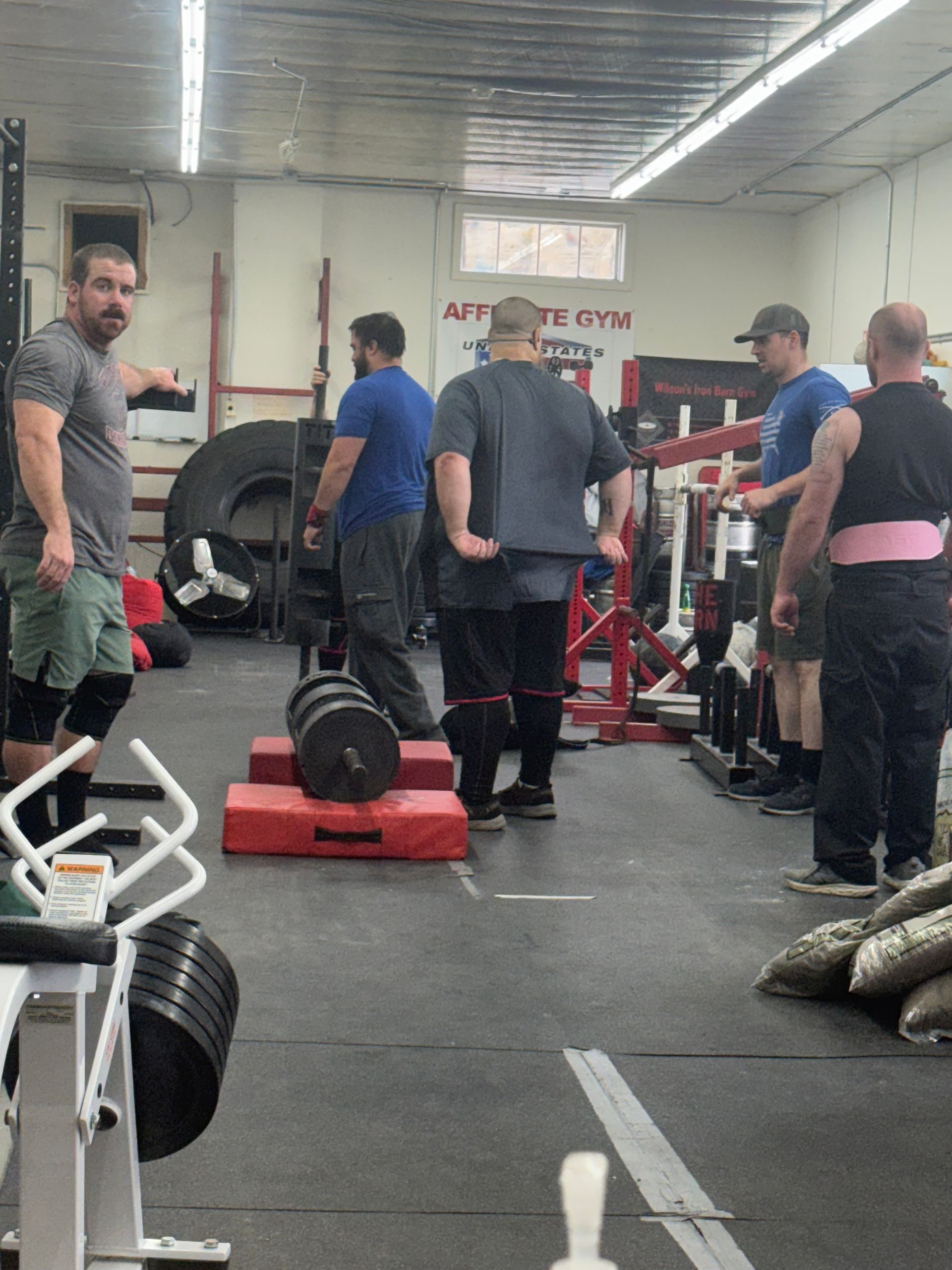 Men in a gym preparing to lift heavy weights. One man points; others watch. A barbell rests on a platform.