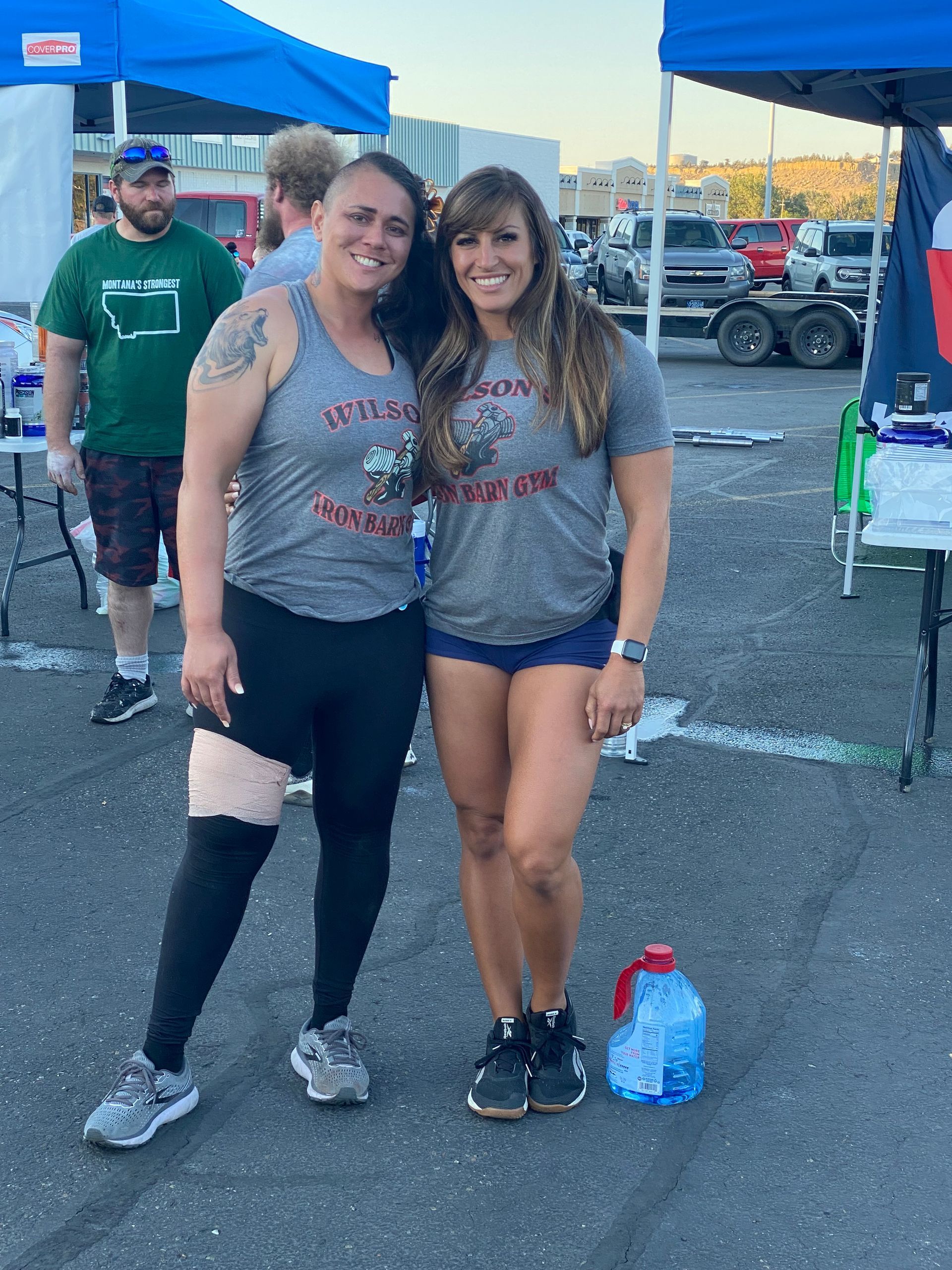Two women in gray shirts pose outdoors, one in leggings, the other in shorts. A man in a green shirt is in the background.