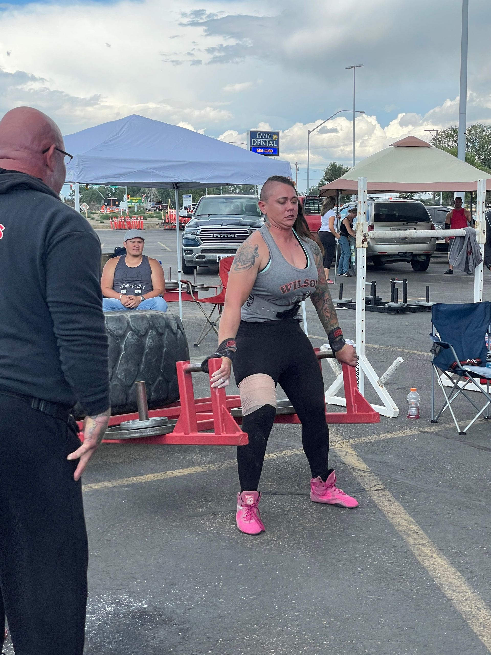 Woman lifting heavy barbell outdoors at a strength competition. Pink shoes, tattoo sleeves, spotter.