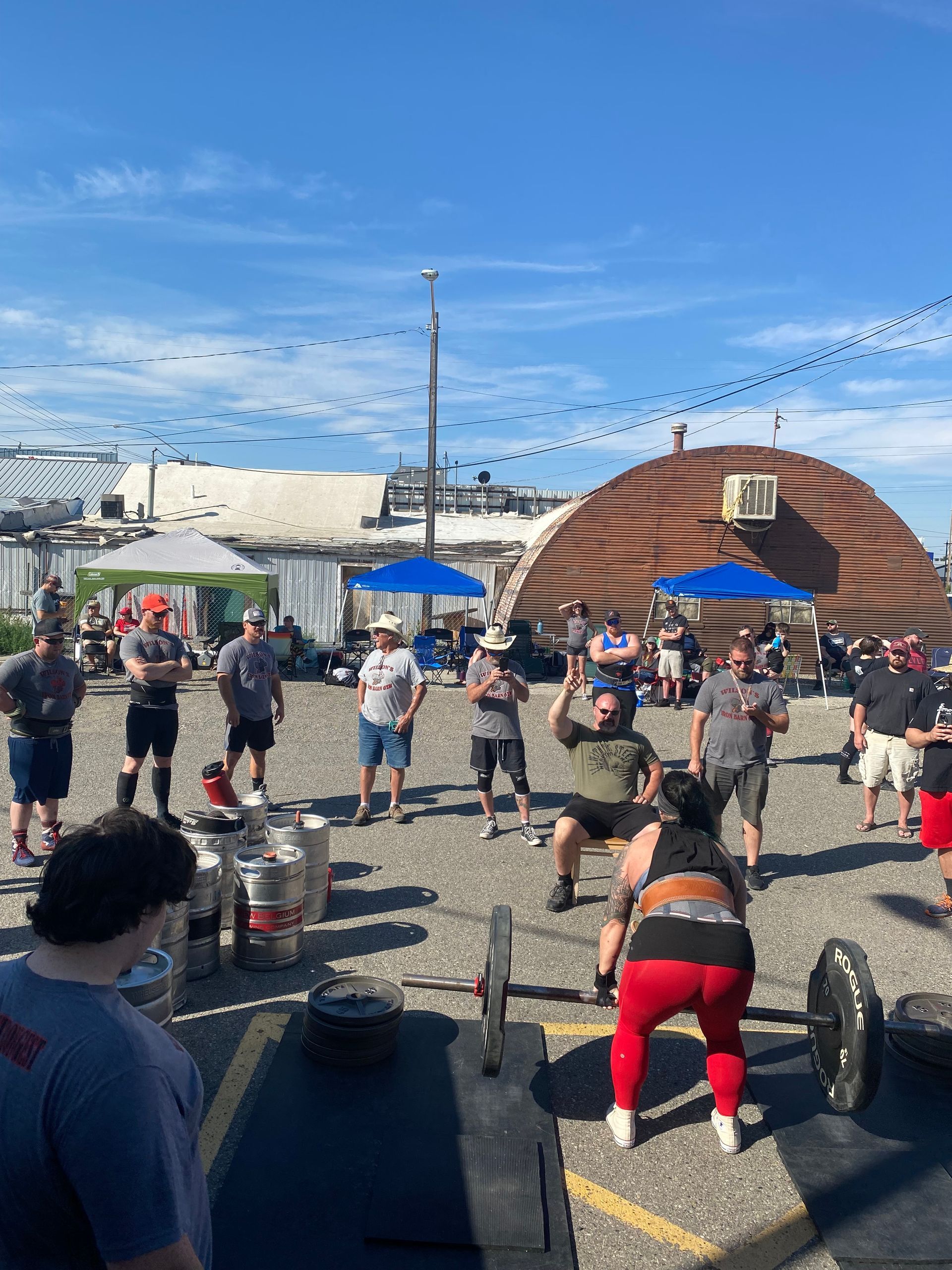 People lifting weights outdoors under a blue sky, with onlookers.