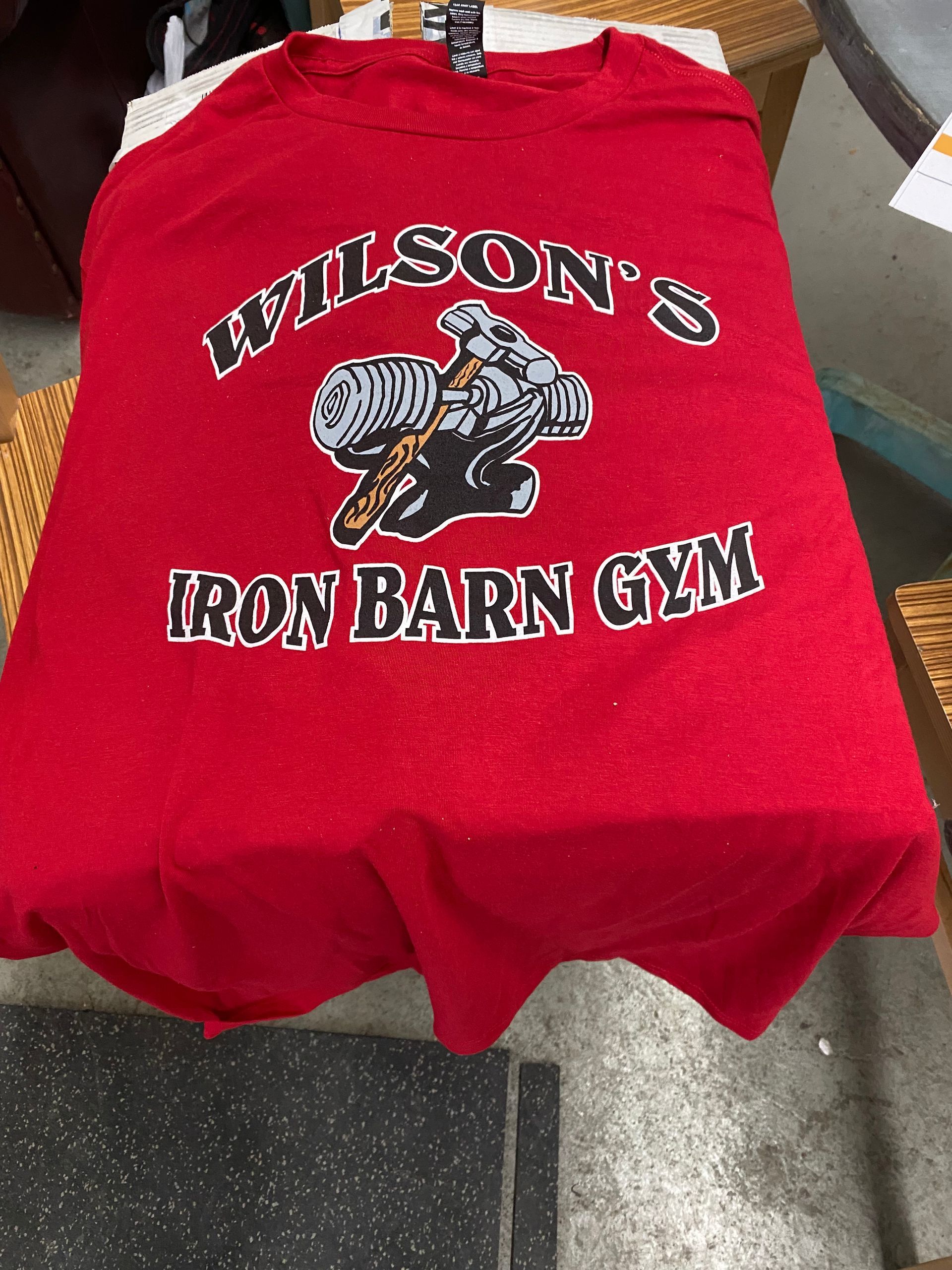 Red Wilson's Iron Barn Gym t-shirt with logo featuring a weightlifter lifting weights, resting on a surface.