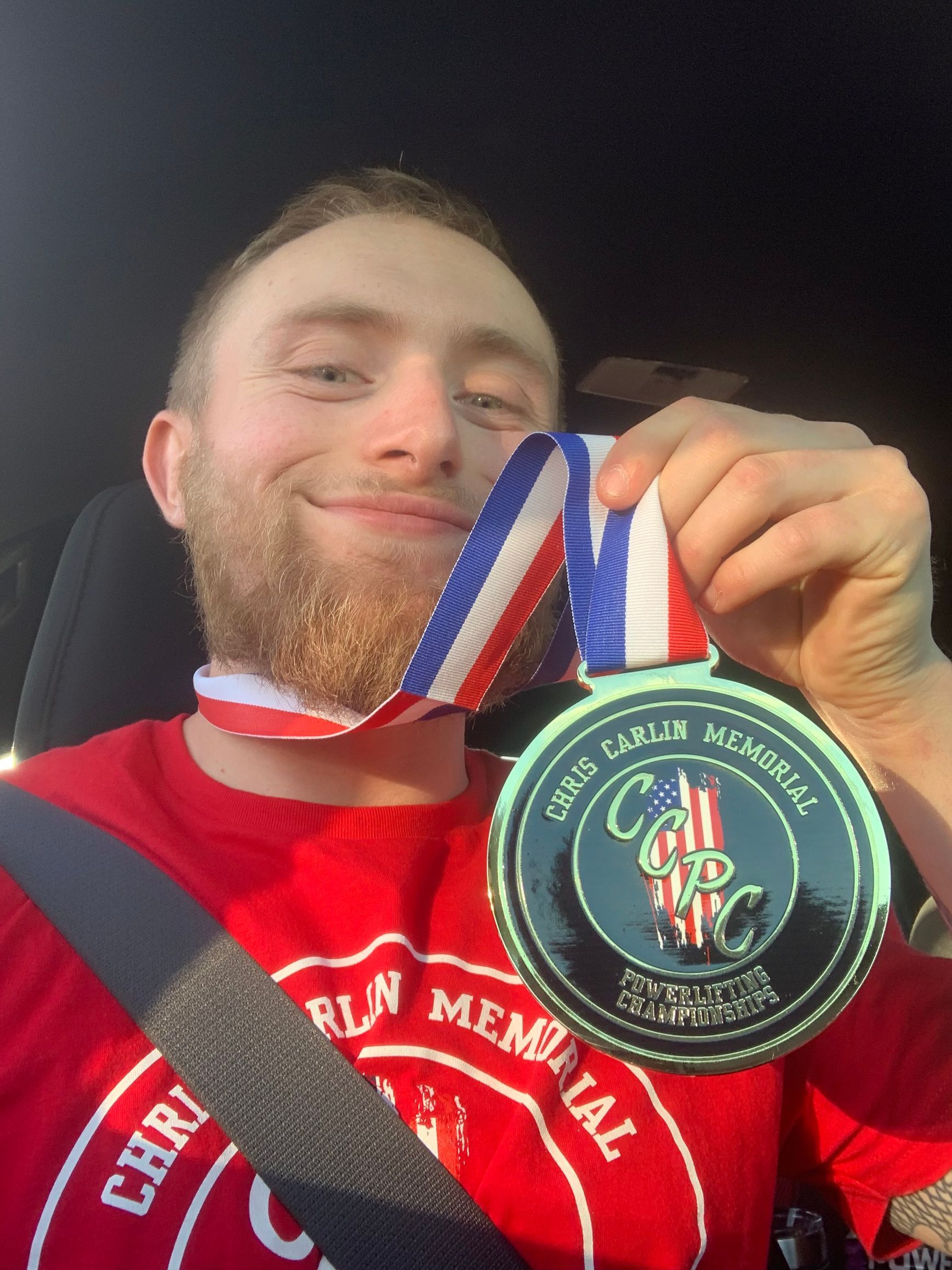 Man in red shirt holding a medal, smiling. The medal has a blue, white, and red ribbon.