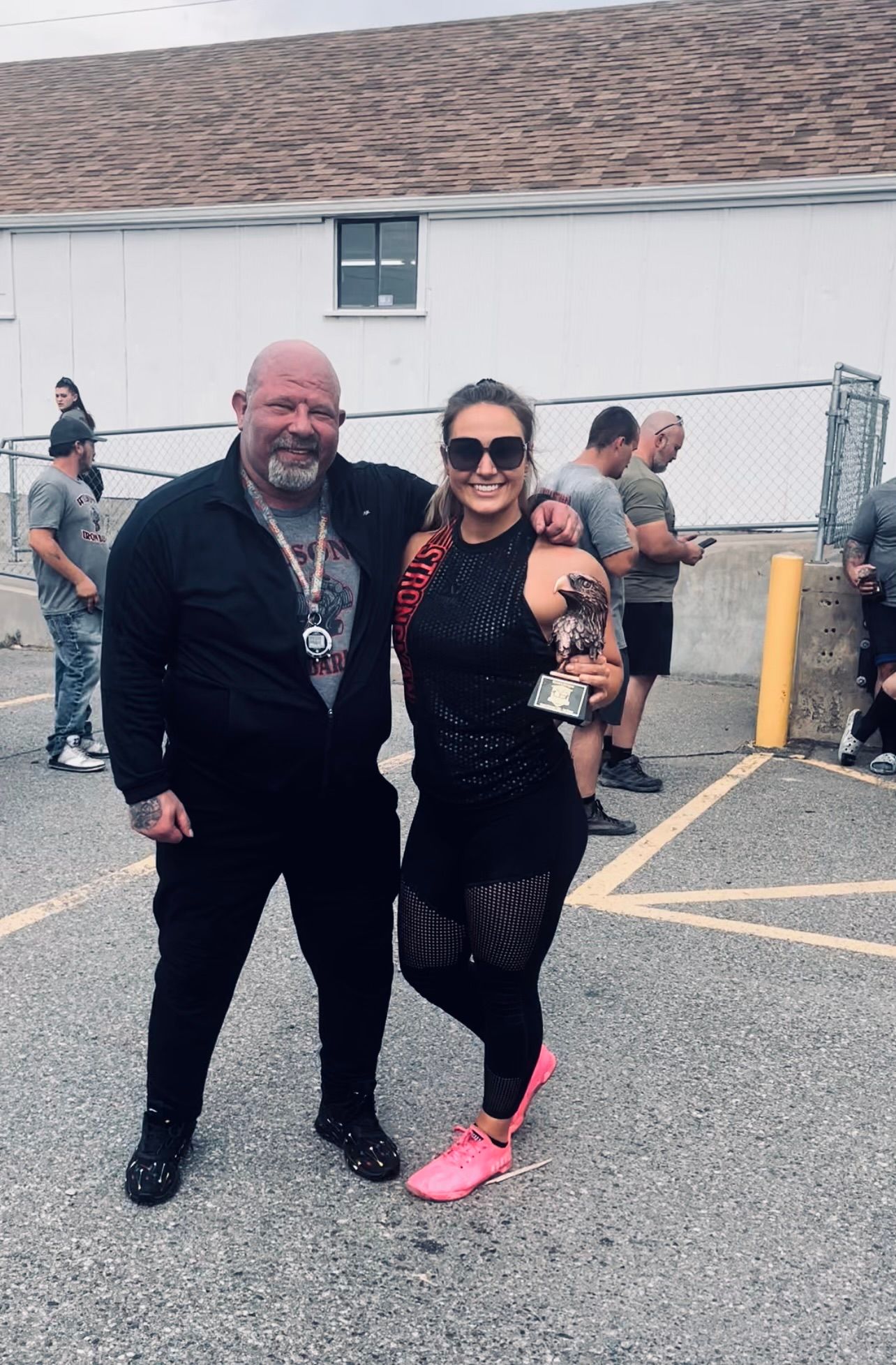 Man and woman posing with a trophy outdoors; man in black outfit, woman in workout gear, pink shoes.