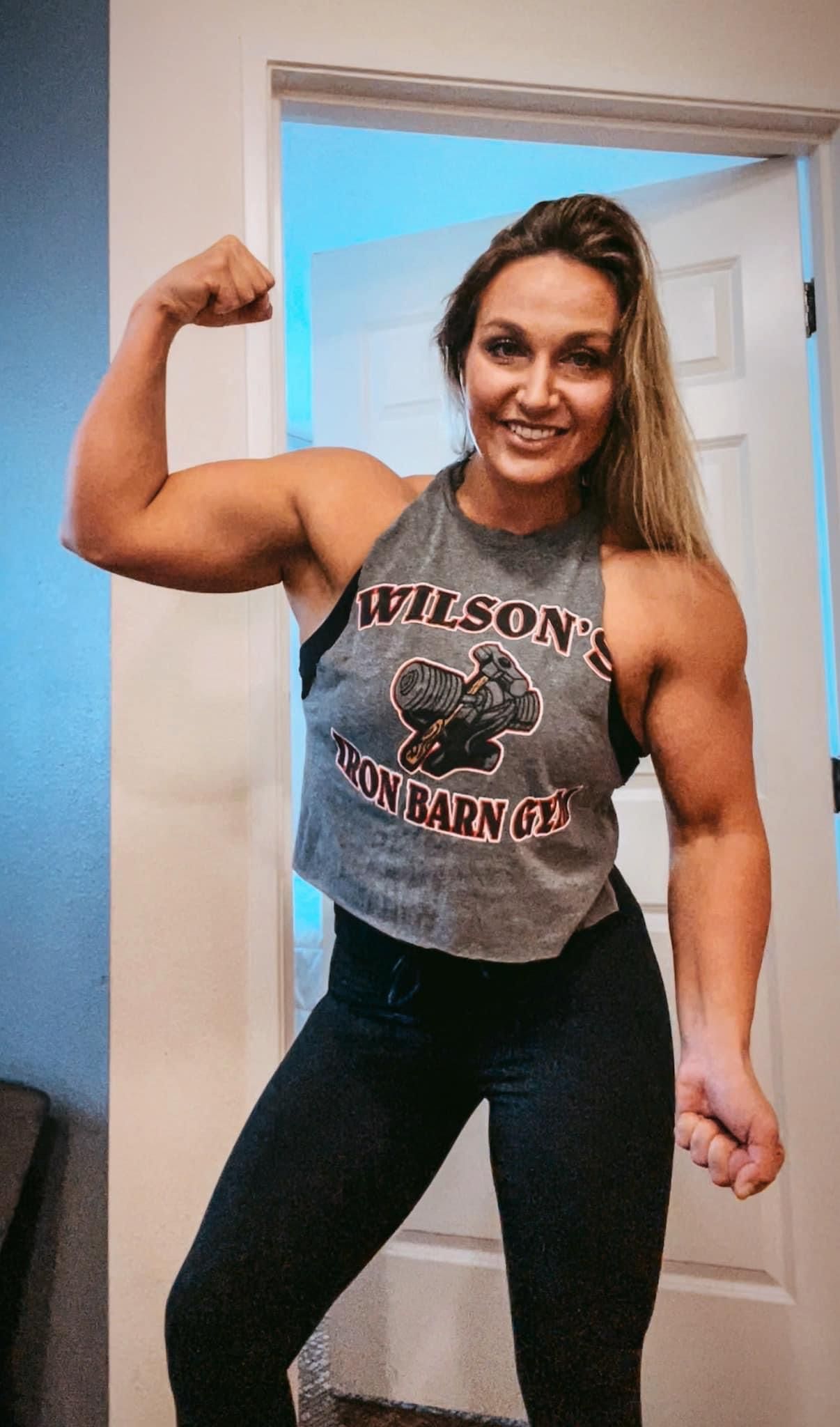 Woman flexing bicep, wearing gray tank and black leggings, smiling. Indoors, by a doorway.