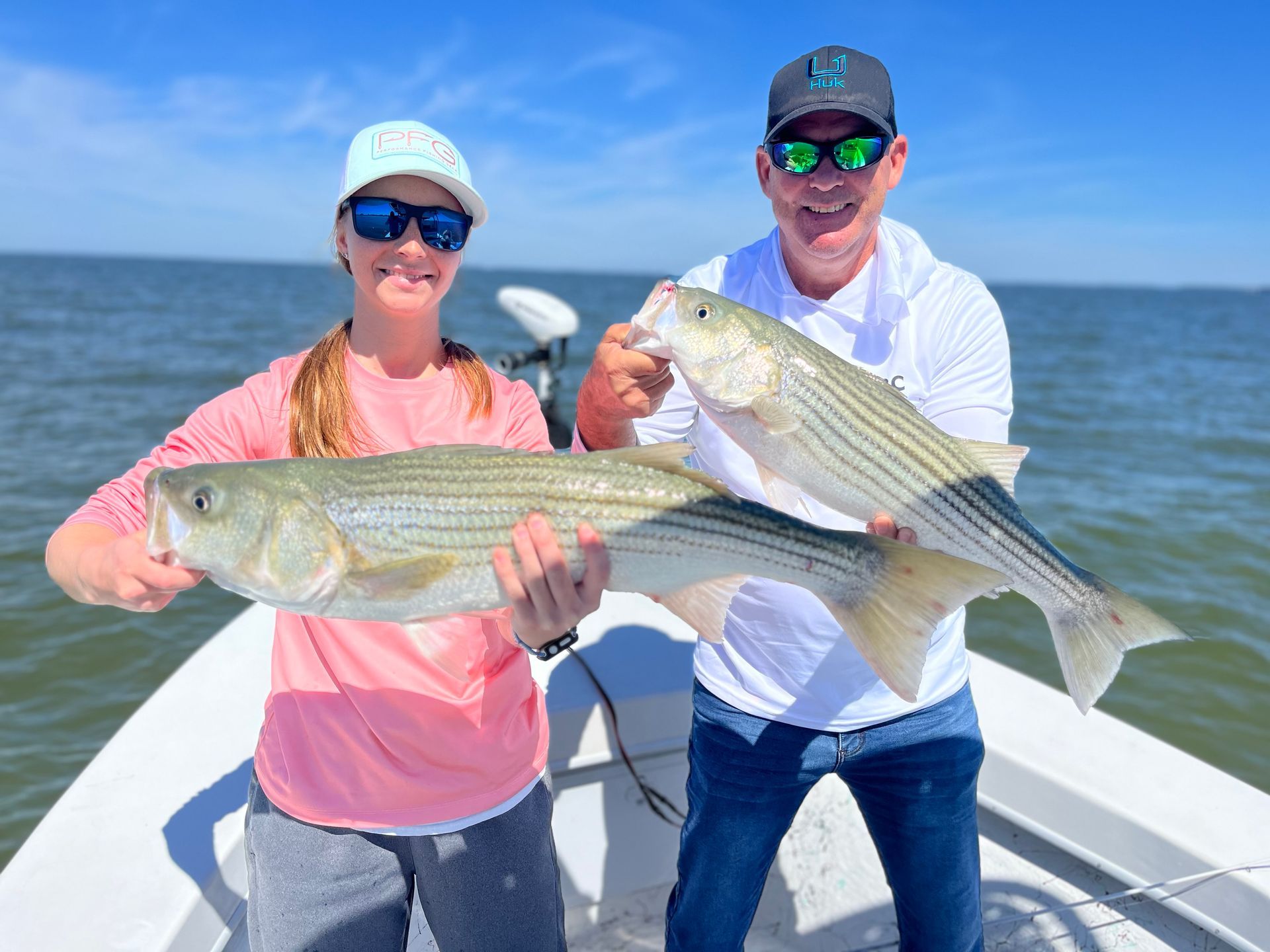 Man on a boat holding a large striped bass fish. Calm water, sunny day.