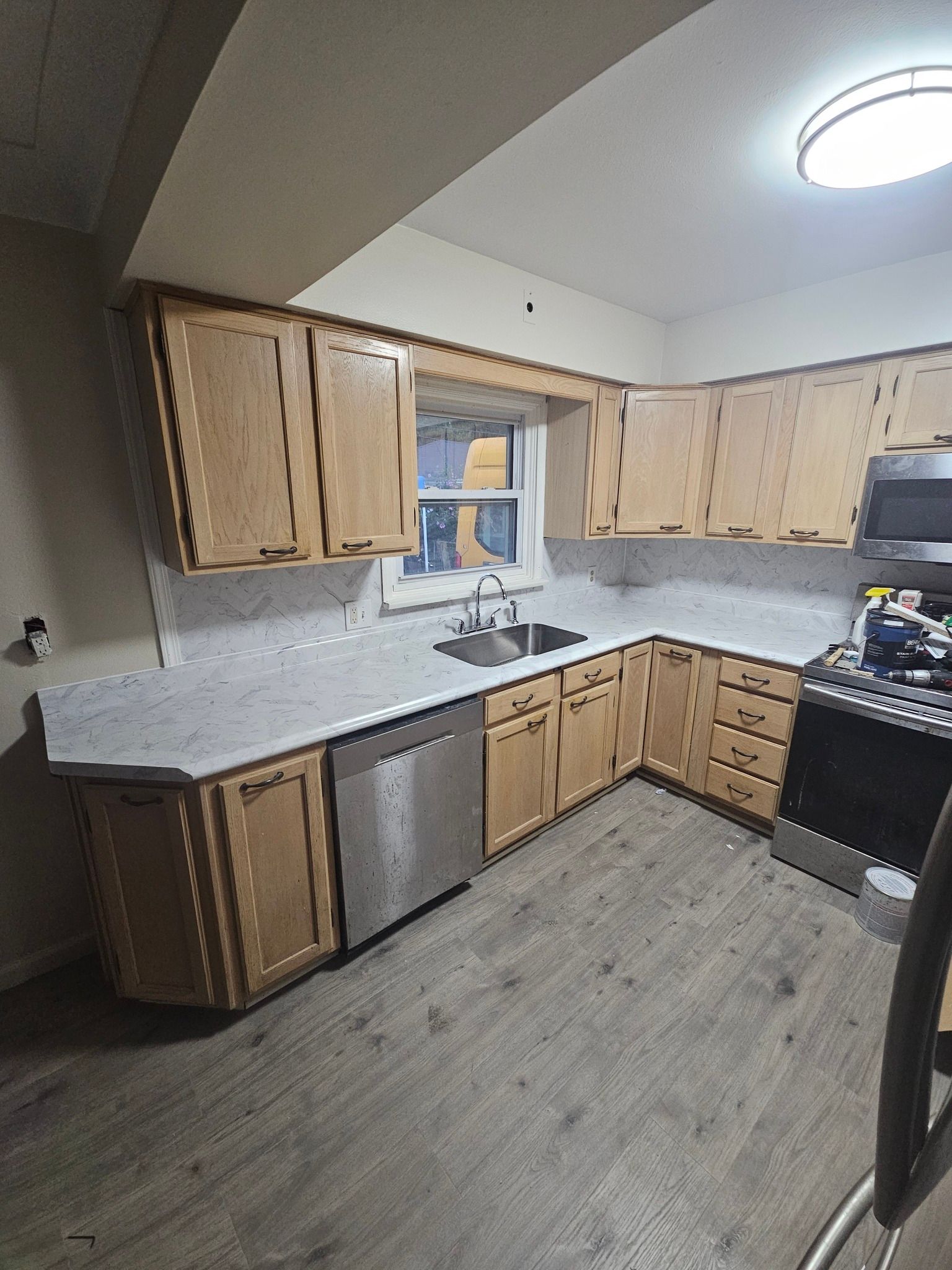 L-shaped kitchen with light brown cabinets, stainless steel appliances, and grey countertops and flooring.