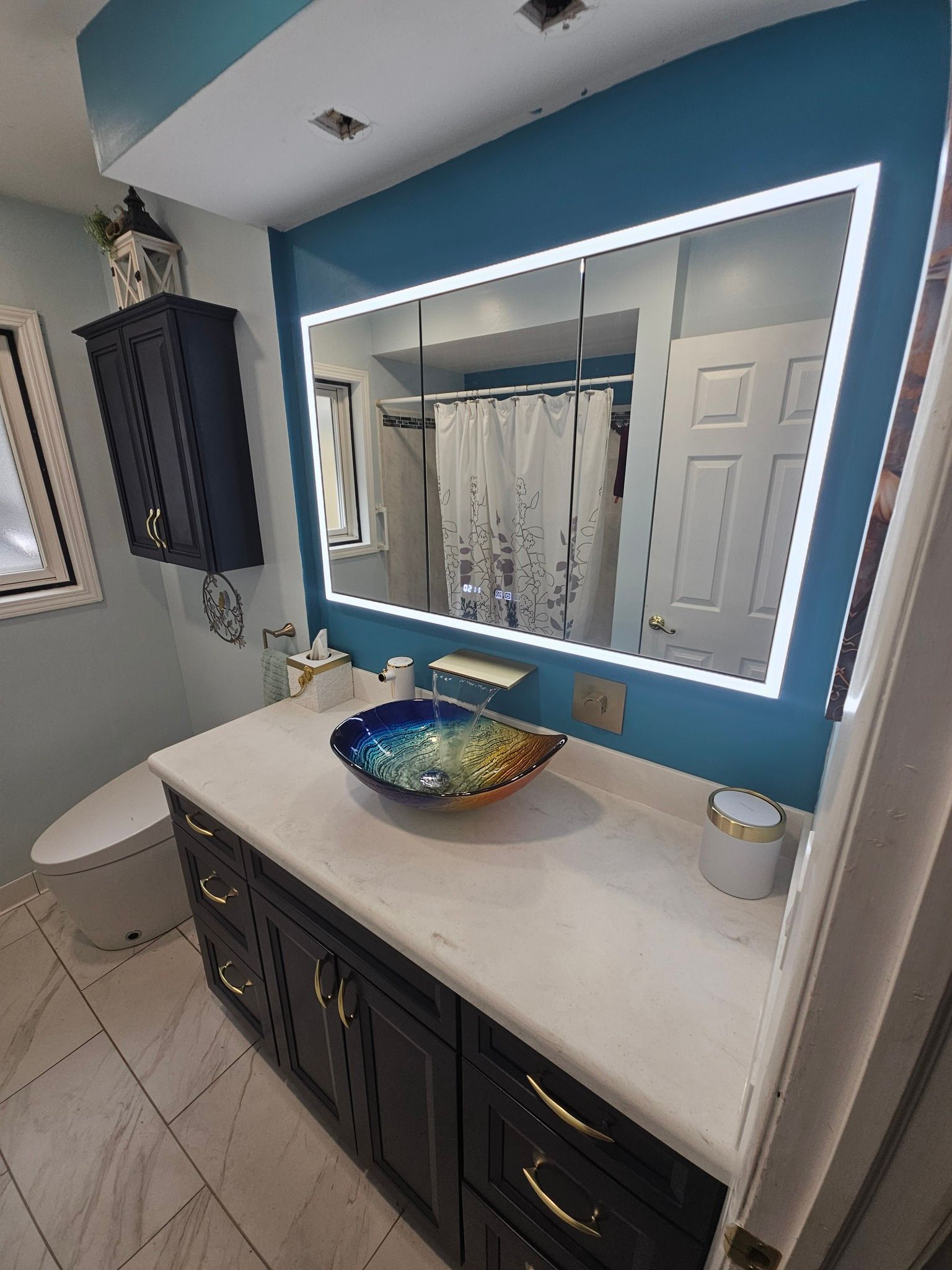 Bathroom with a blue wall, large illuminated mirror, dark vanity, and a colorful sink bowl.