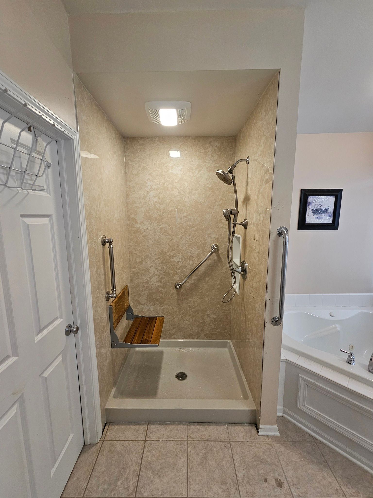 Accessible shower with fold-down seat, grab bars, and hand shower. Tan walls, cream ceiling, and a tiled floor.