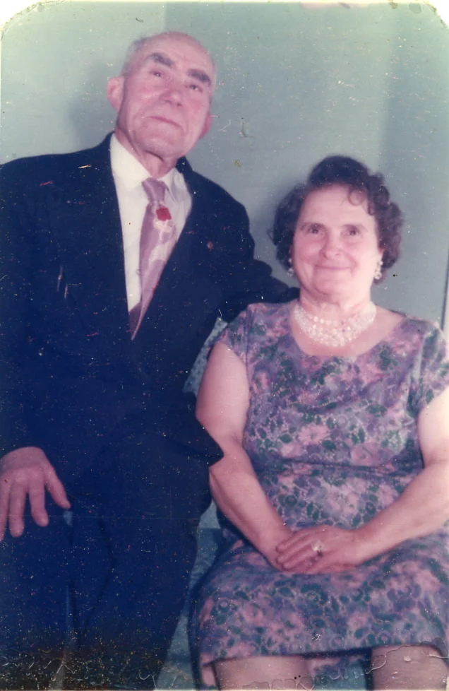Elderly couple posing together. Man in suit, woman in floral dress, seated.