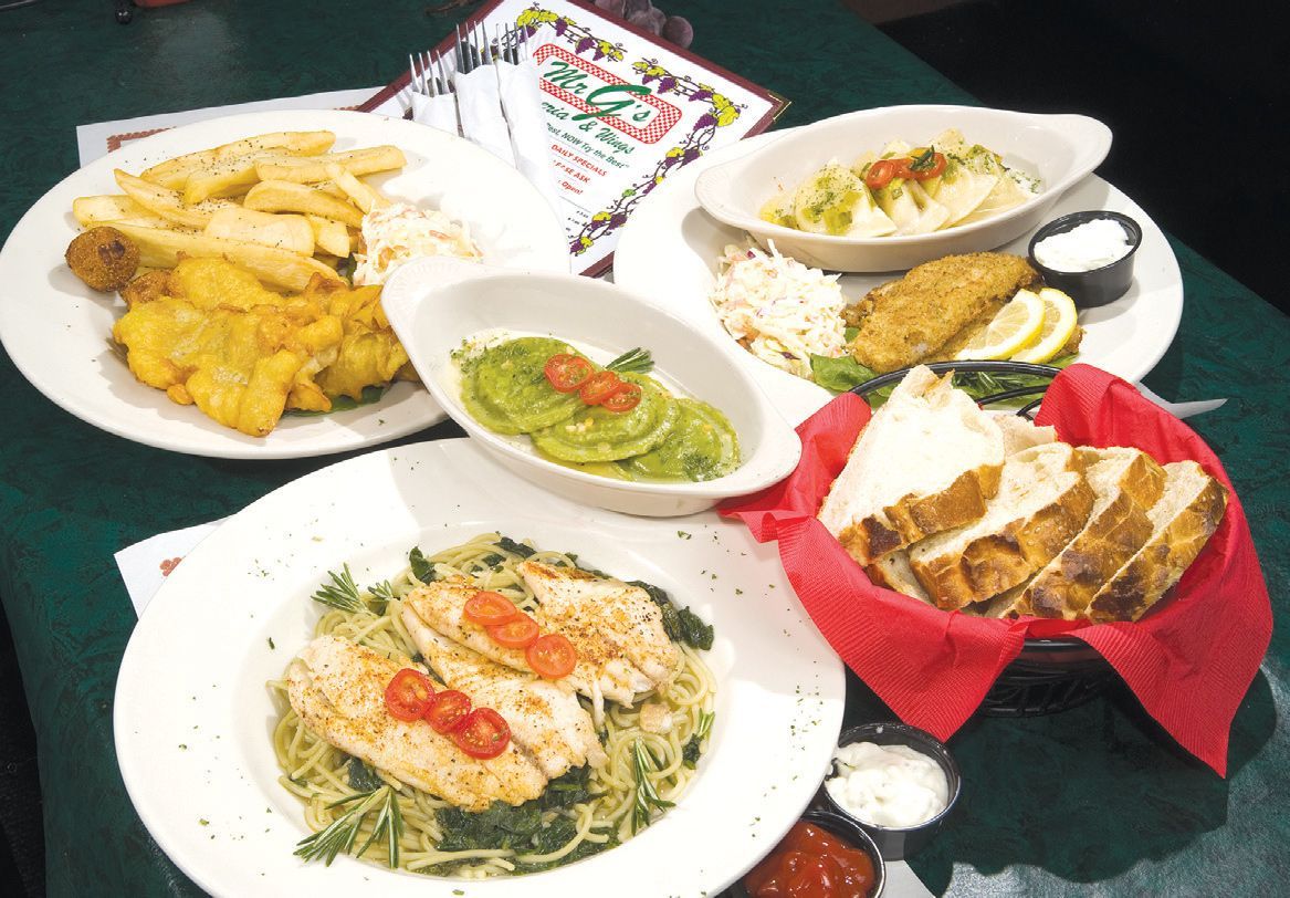Plates of food, including pasta, fish, fries, and avocado, on a green table. Bread and sauces are also present.