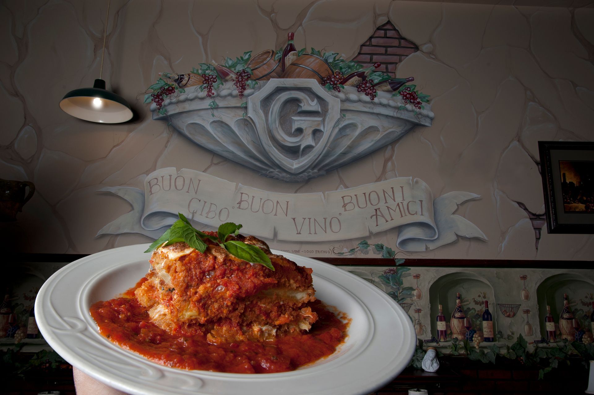 Lasagna on a white plate in front of a painted Italian restaurant mural.