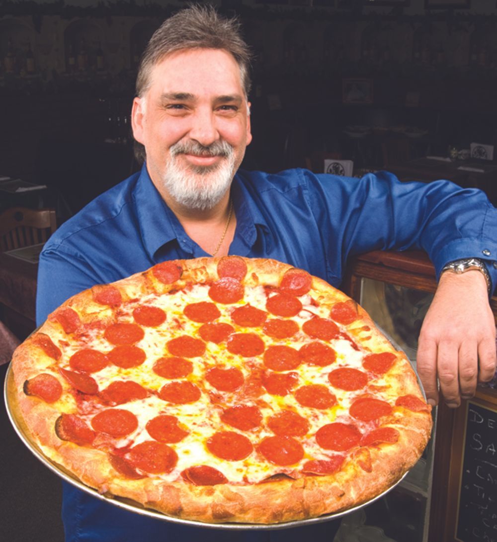 Man holding a large pepperoni pizza, smiling. He wears a blue shirt; pizza on a metal tray.