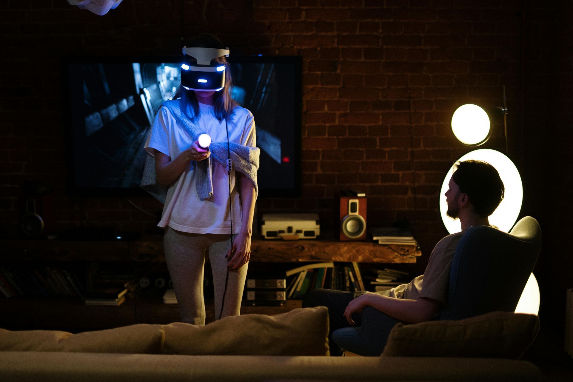 A woman wearing a virtual reality headset is playing a video game while a man sits on a couch.