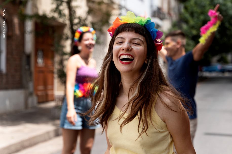 Mulher com coroa de arco-íris e expressão alegre, celebrando ao ar livre com outras pessoas.