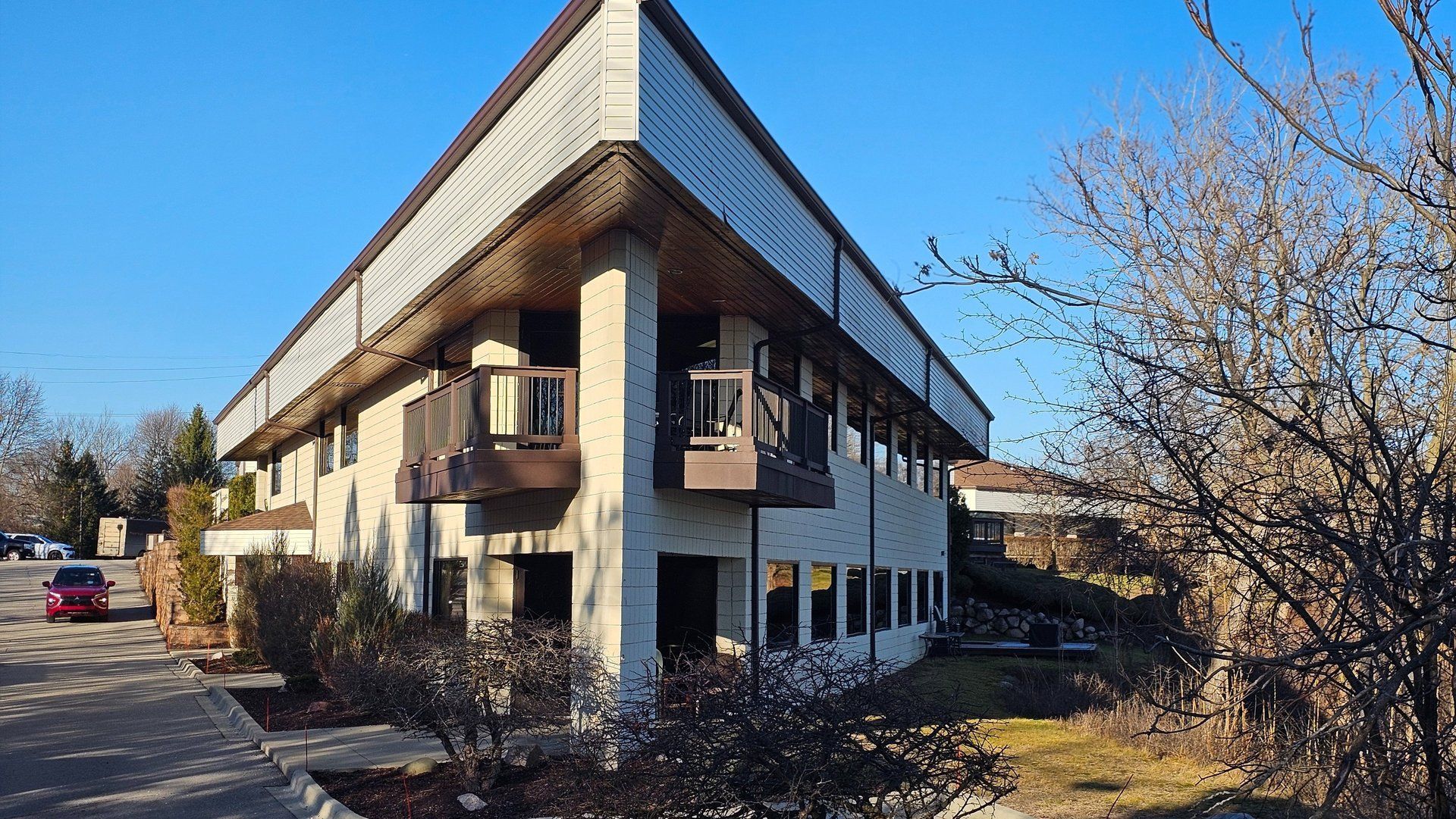 Two-story building with tan exterior, balconies, and large overhanging roof. Blue sky and trees in the background.